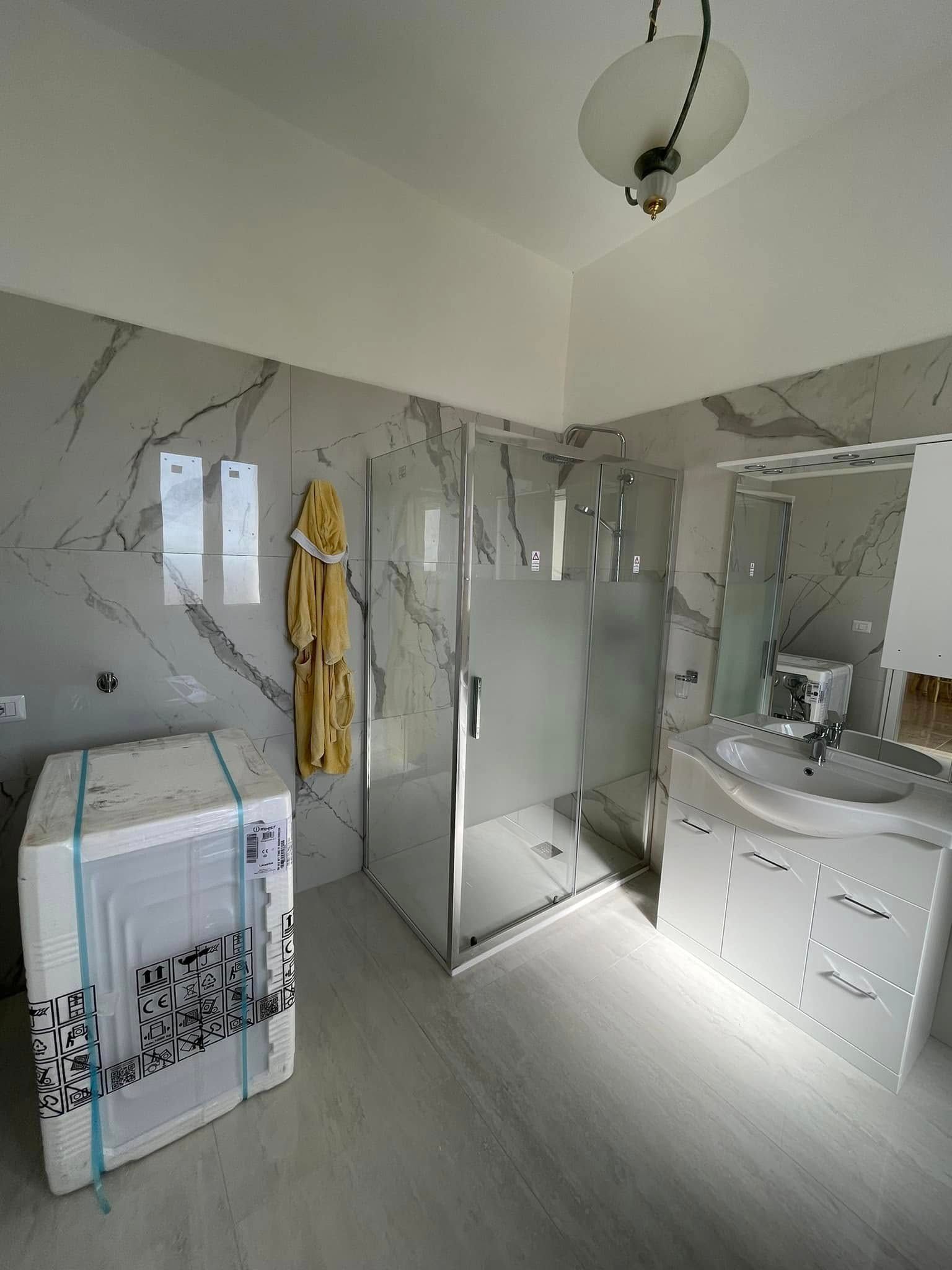 Bagno luminoso con piastrelle effetto marmo, doccia, mobiletto sotto il lavabo e lavatrice vicino a una finestra.