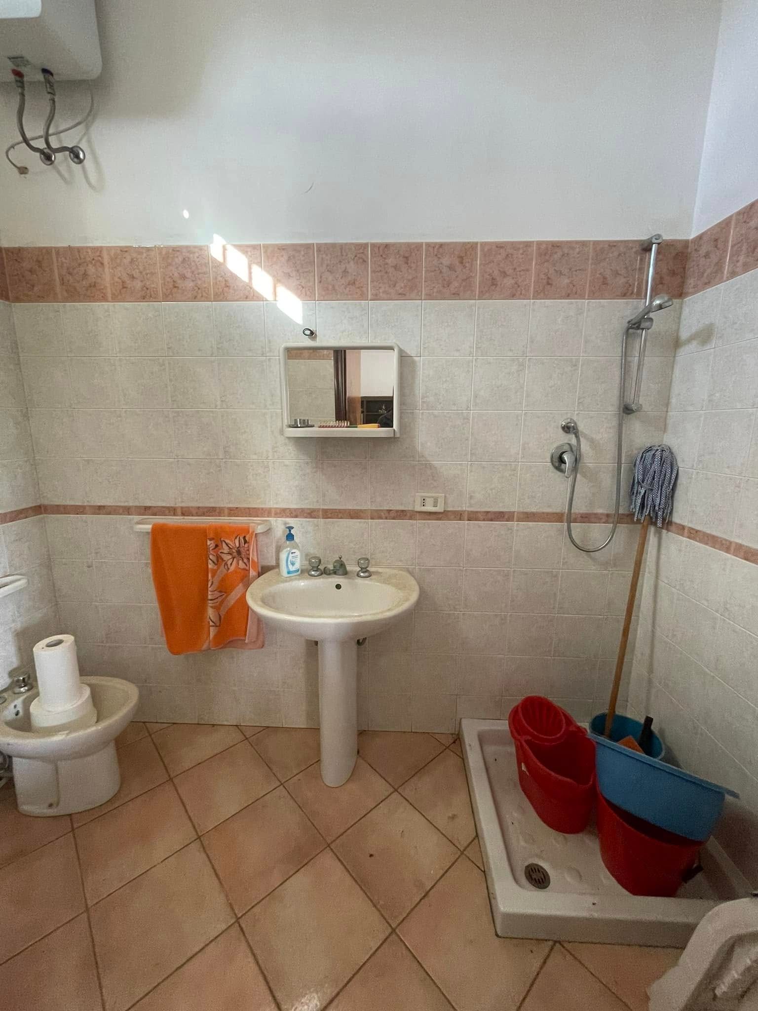 Piccolo bagno piastrellato con lavandino, WC, doccia, asciugamano arancione e secchi rossi e blu sul pavimento.