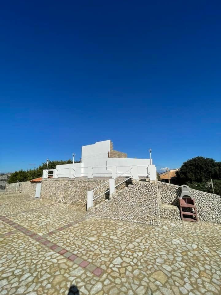 Edificio bianco su una terrazza in pietra sotto un cielo azzurro limpido, con scalini e uno sfondo roccioso.