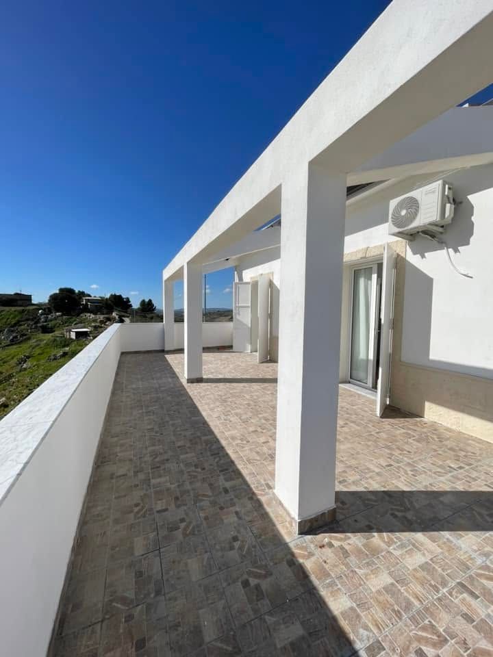 Terrazza panoramica soleggiata con colonne bianche, pavimentazione in pietra e cielo azzurro su un paesaggio erboso.