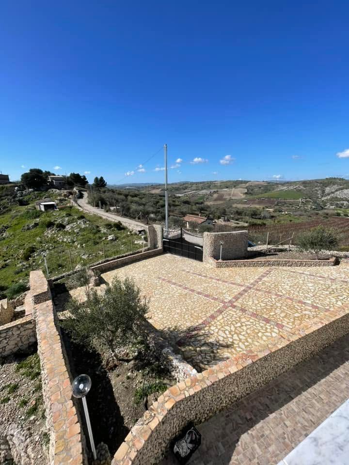 Terrazza rurale in collina con muri in pietra, sentiero di ghiaia e cielo azzurro sopra campi ondulati.