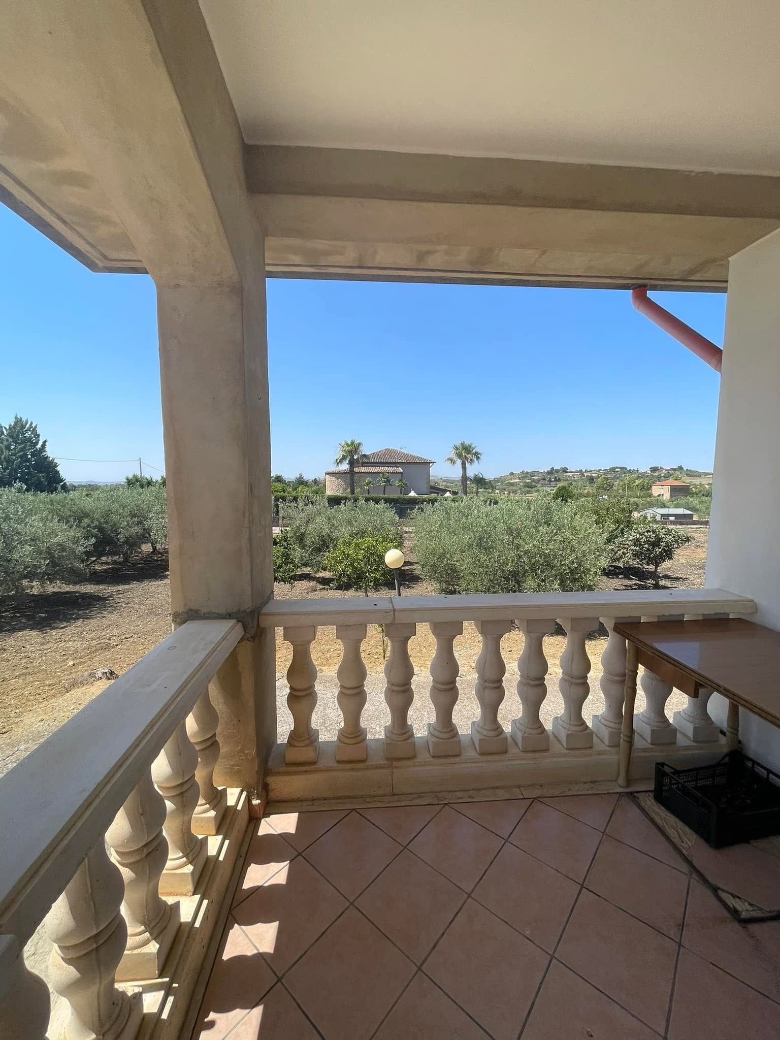 Vista dal portico sulla campagna soleggiata con balaustra bianca, pavimento piastrellato e alberi in lontananza sotto un cielo azzurro.