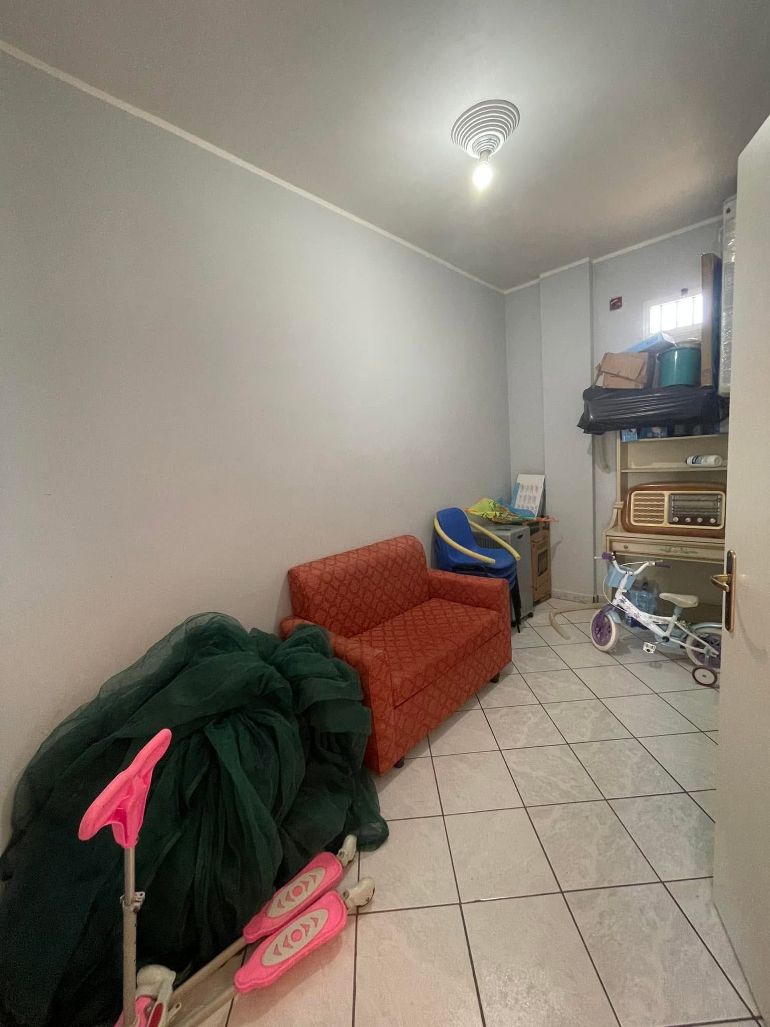 Stretto corridoio con divano rosso, coperta verde, giocattolo rosa e scaffali ingombri sotto una lampada a soffitto.