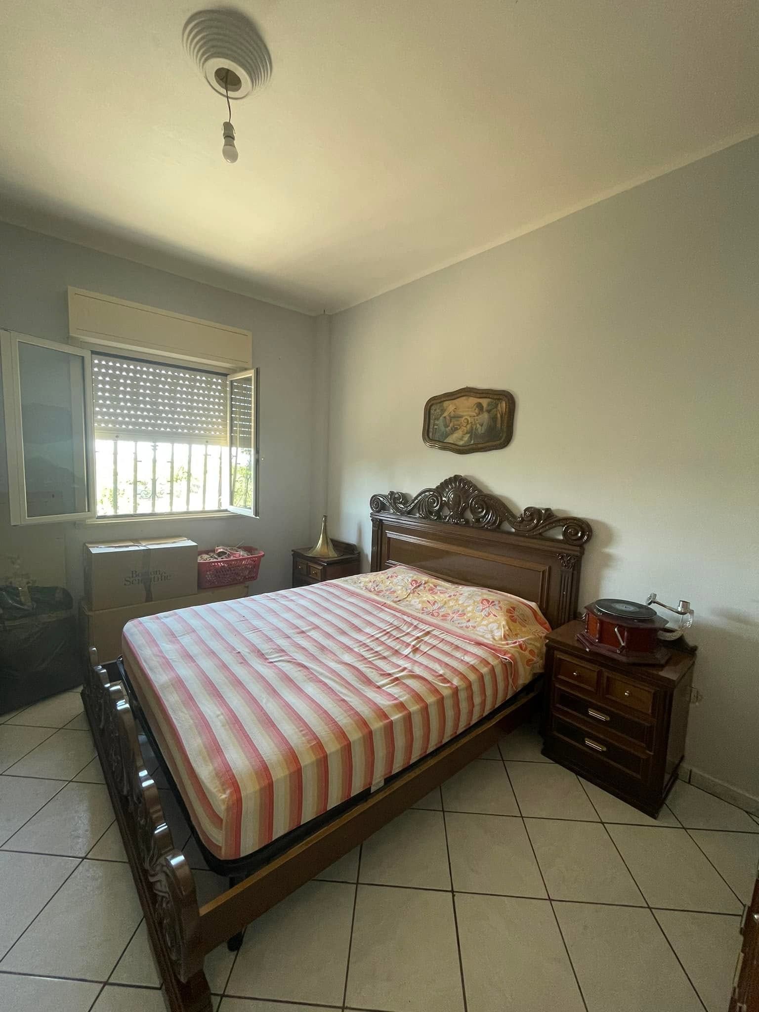 Piccola camera da letto con letto in legno intagliato, coperta a righe, comodino, finestra e pavimento piastrellato.
