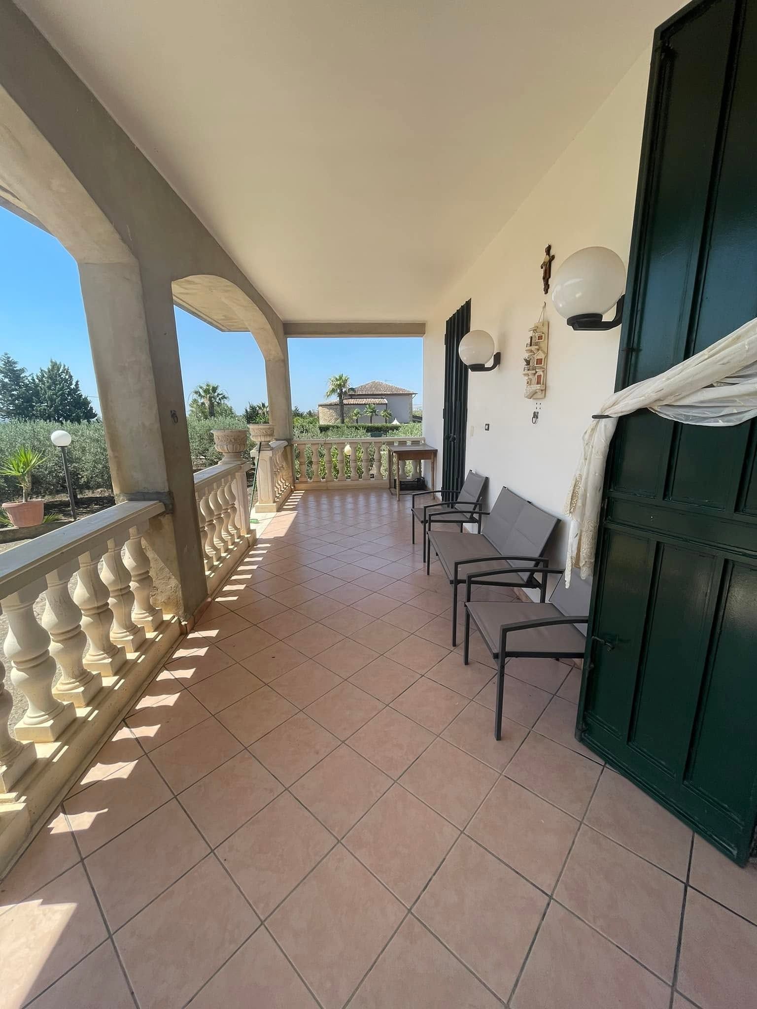 Veranda soleggiata e coperta con pavimento piastrellato, ringhiera bianca, sedie nere e persiane verdi che si affaccia su un giardino.