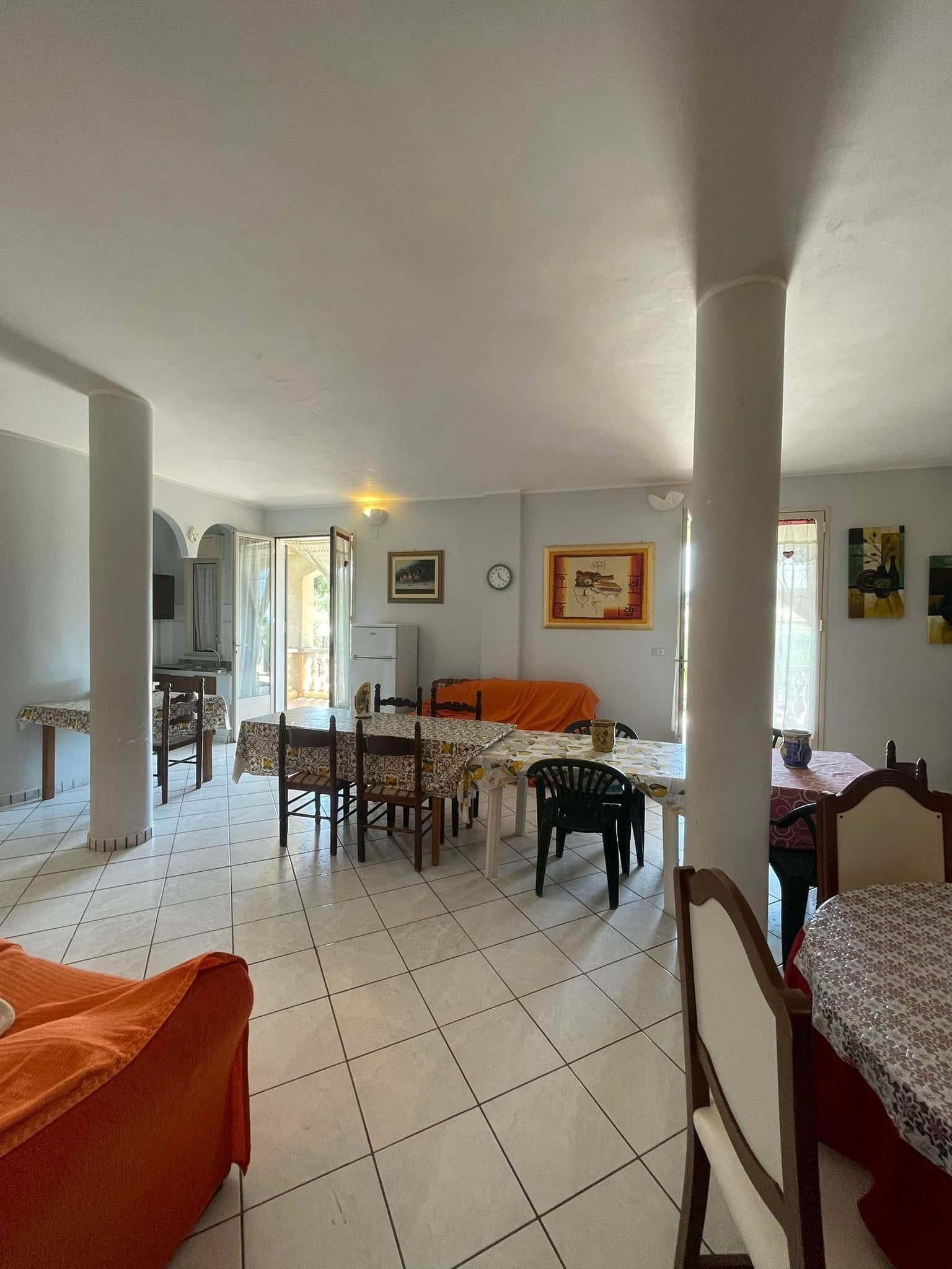 Luminoso soggiorno piastrellato con divani arancioni, tavolo da pranzo e porta aperta su un patio.