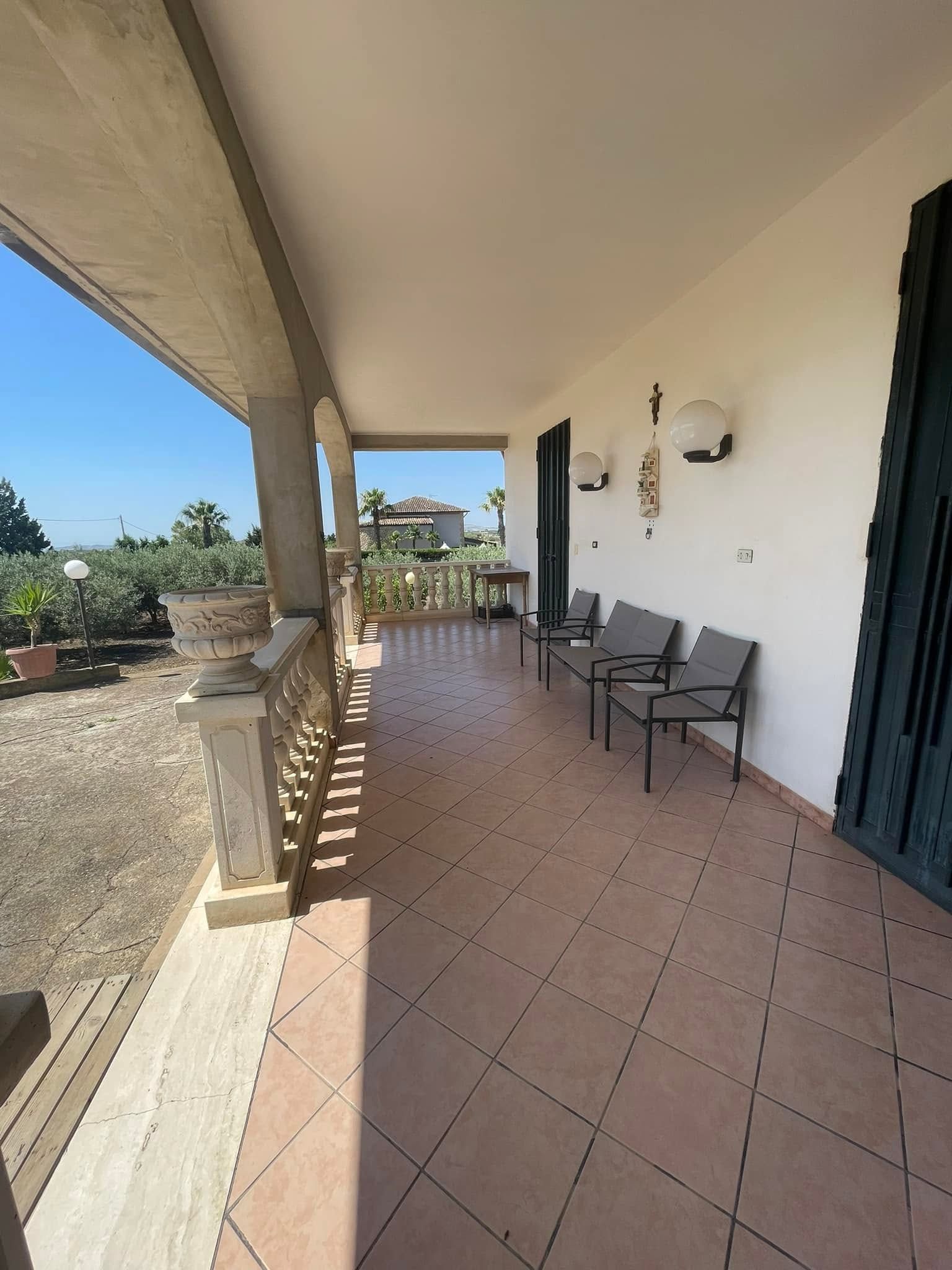 Veranda coperta con pavimento piastrellato, sedie accanto a una parete bianca e vista sul verde esterno.