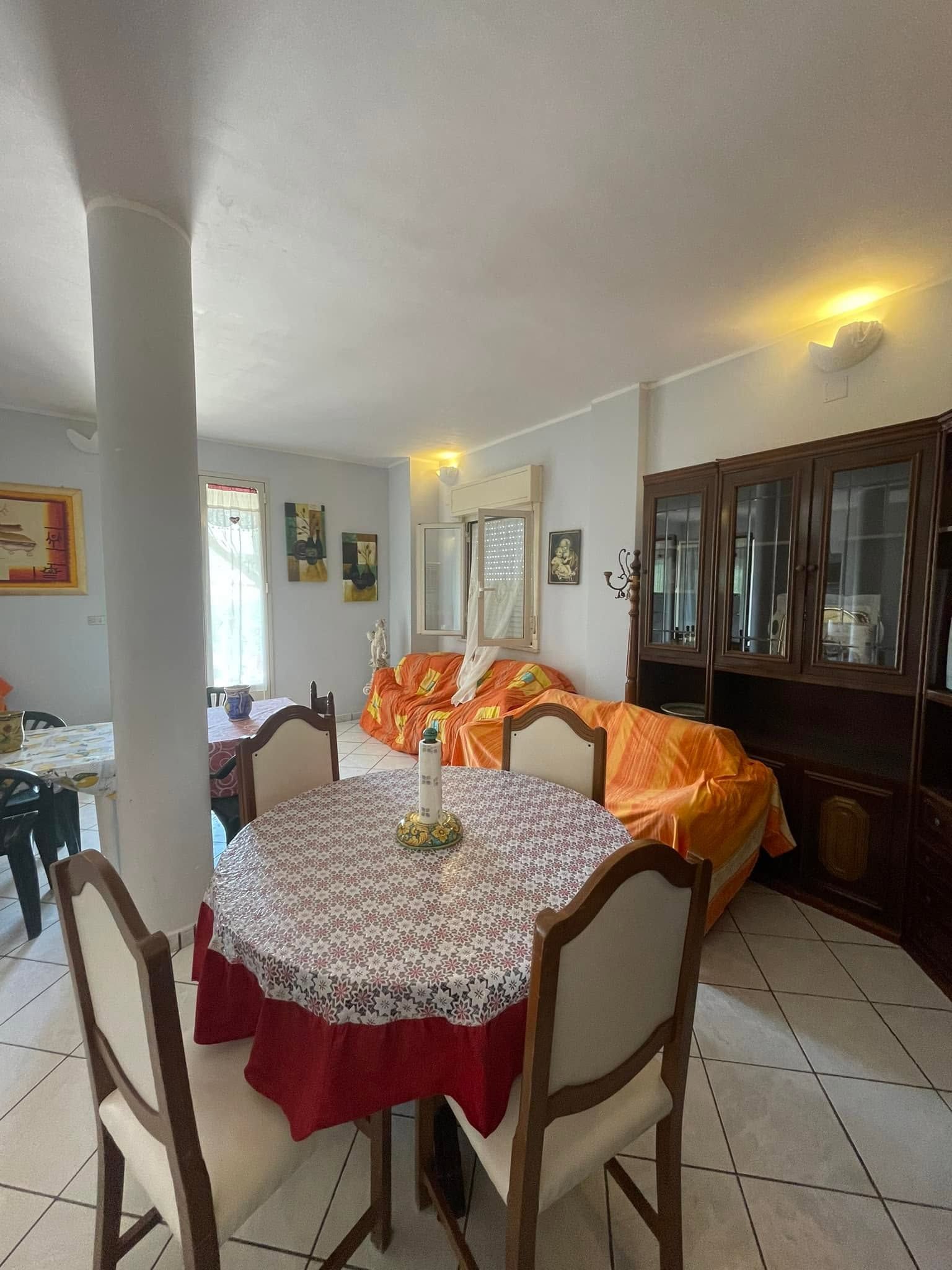 Sala da pranzo luminosa con tavolo apparecchiato, divano arancione e mobile in legno scuro su pavimento piastrellato.