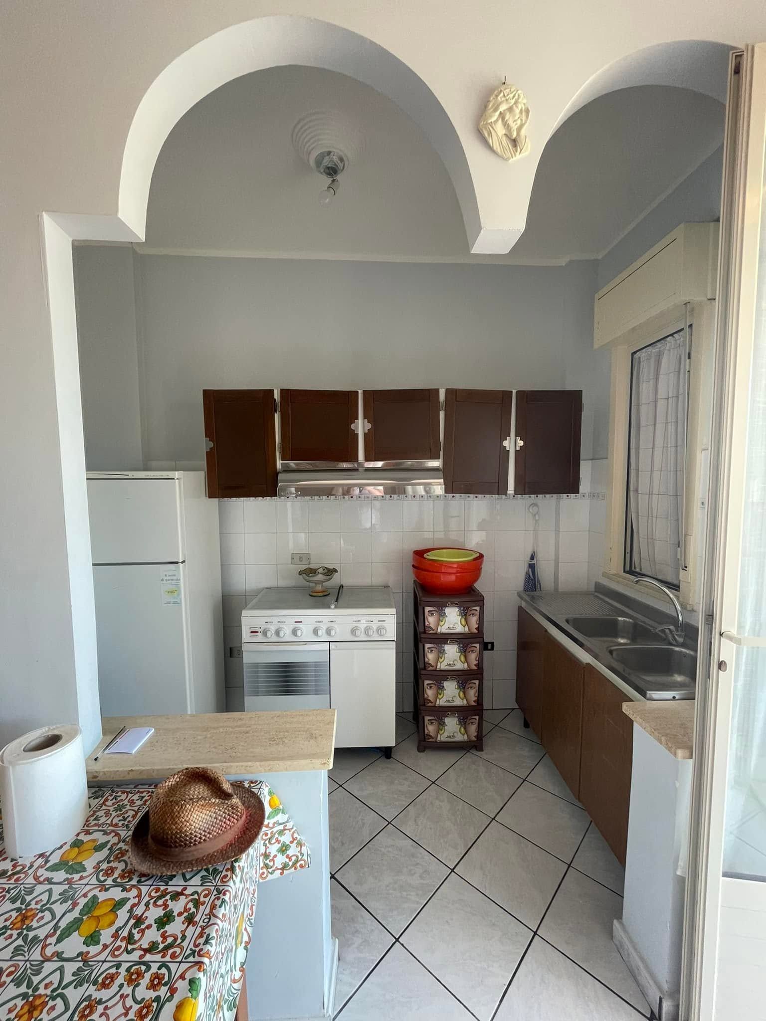 Piccola cucina con aperture ad arco, pareti grigie, mobili scuri, fornelli, lavello e un tavolo in primo piano.
