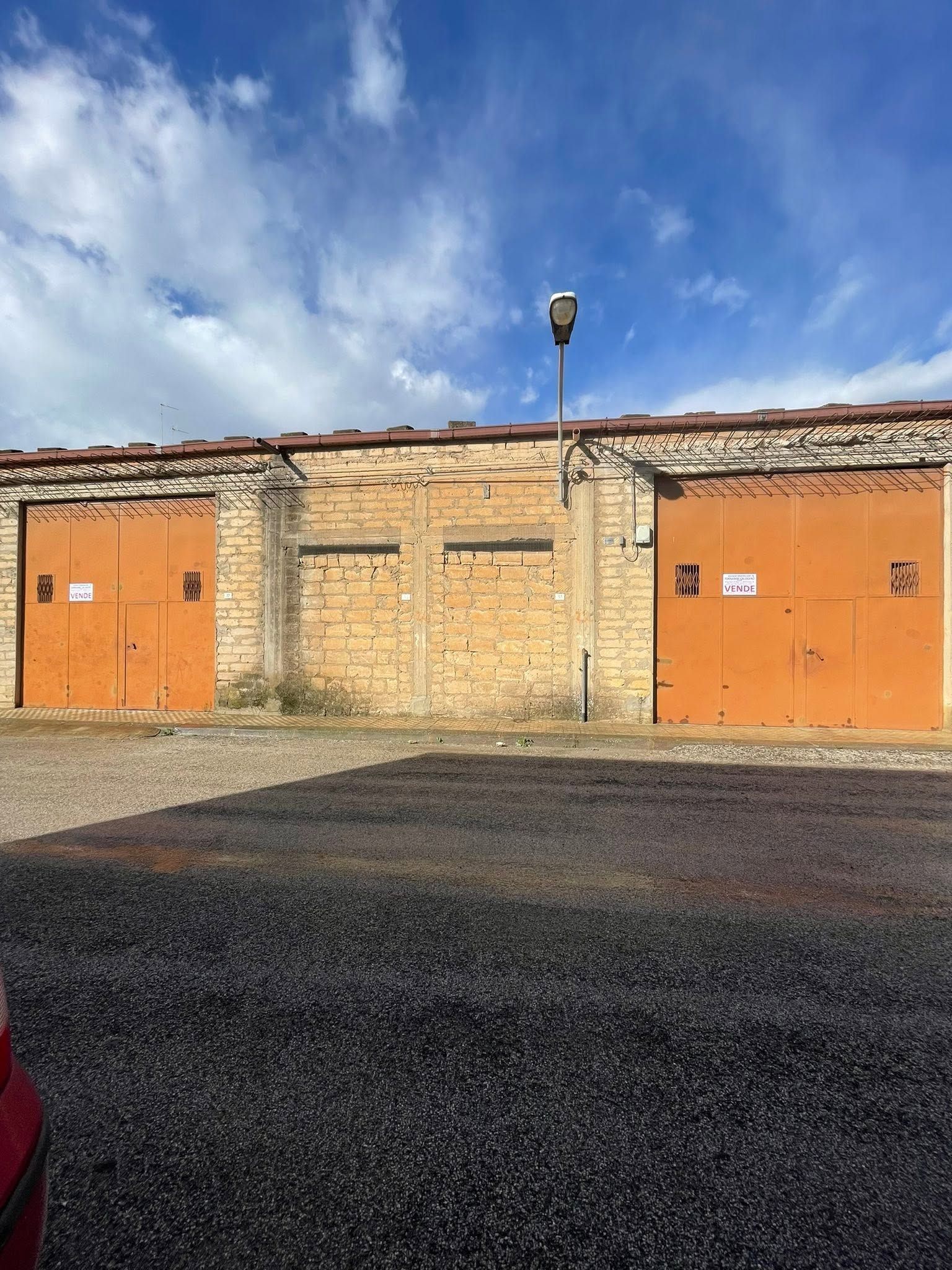 Edificio in mattoni con due porte arancioni e un cielo azzurro nuvoloso sopra un lotto di asfalto vuoto.