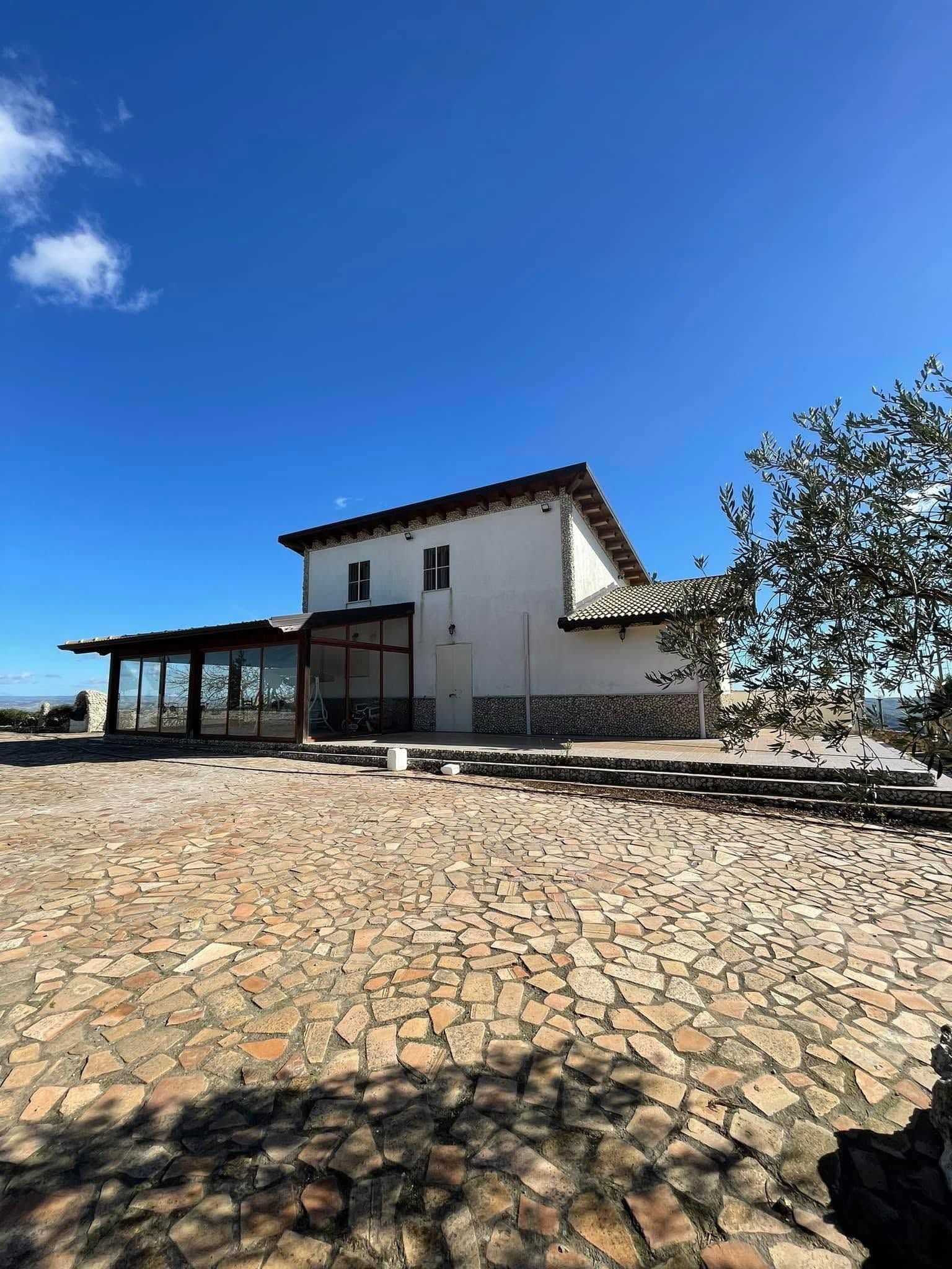 Cortile in pietra con una casa rustica bianca sotto un cielo azzurro brillante