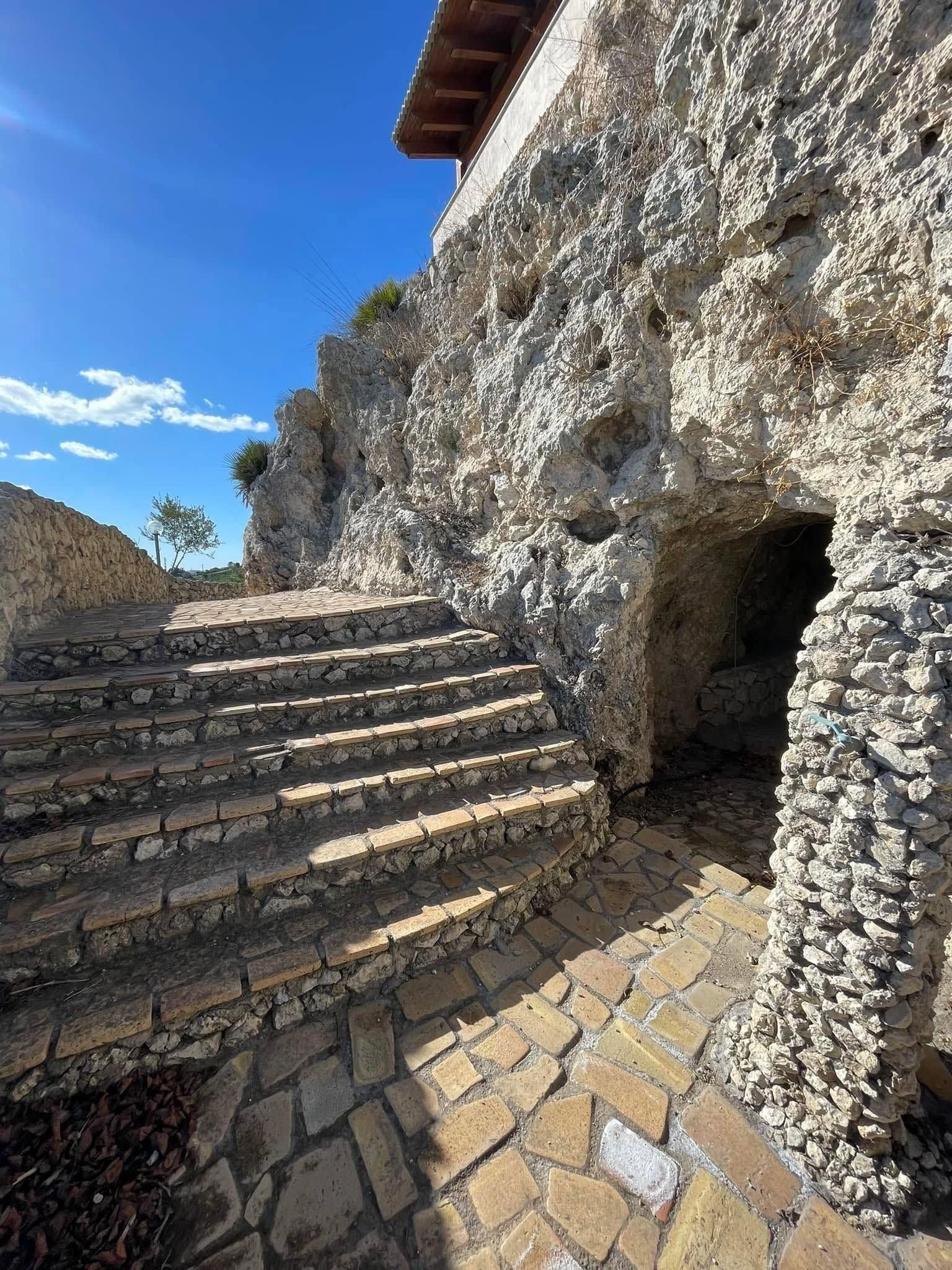 Scalini di pietra accanto a una scogliera rocciosa e un'apertura simile a una grotta sotto un cielo azzurro brillante