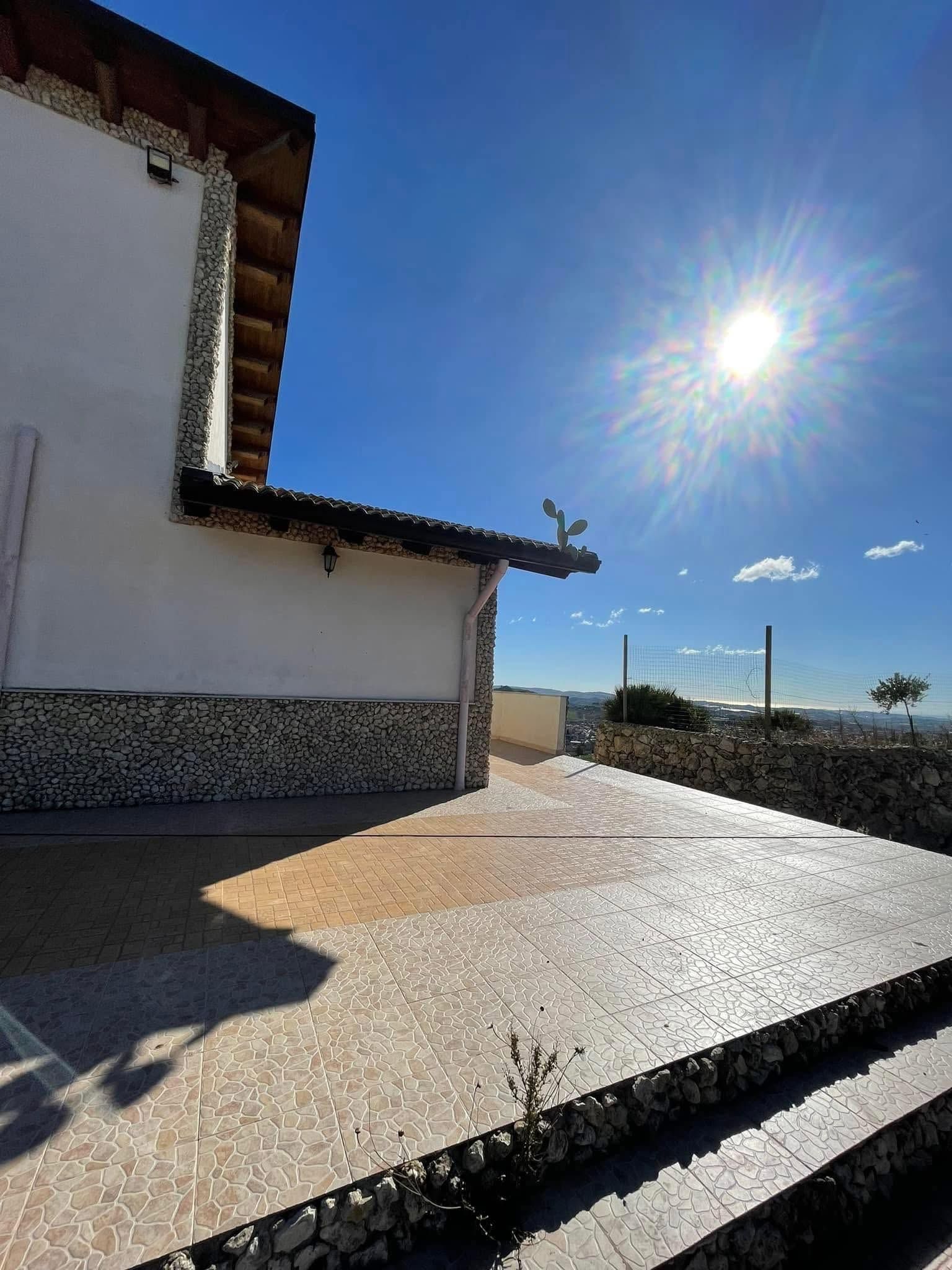 Patio soleggiato accanto a una casa bianca, con scalini in pietra e un cielo azzurro brillante.
