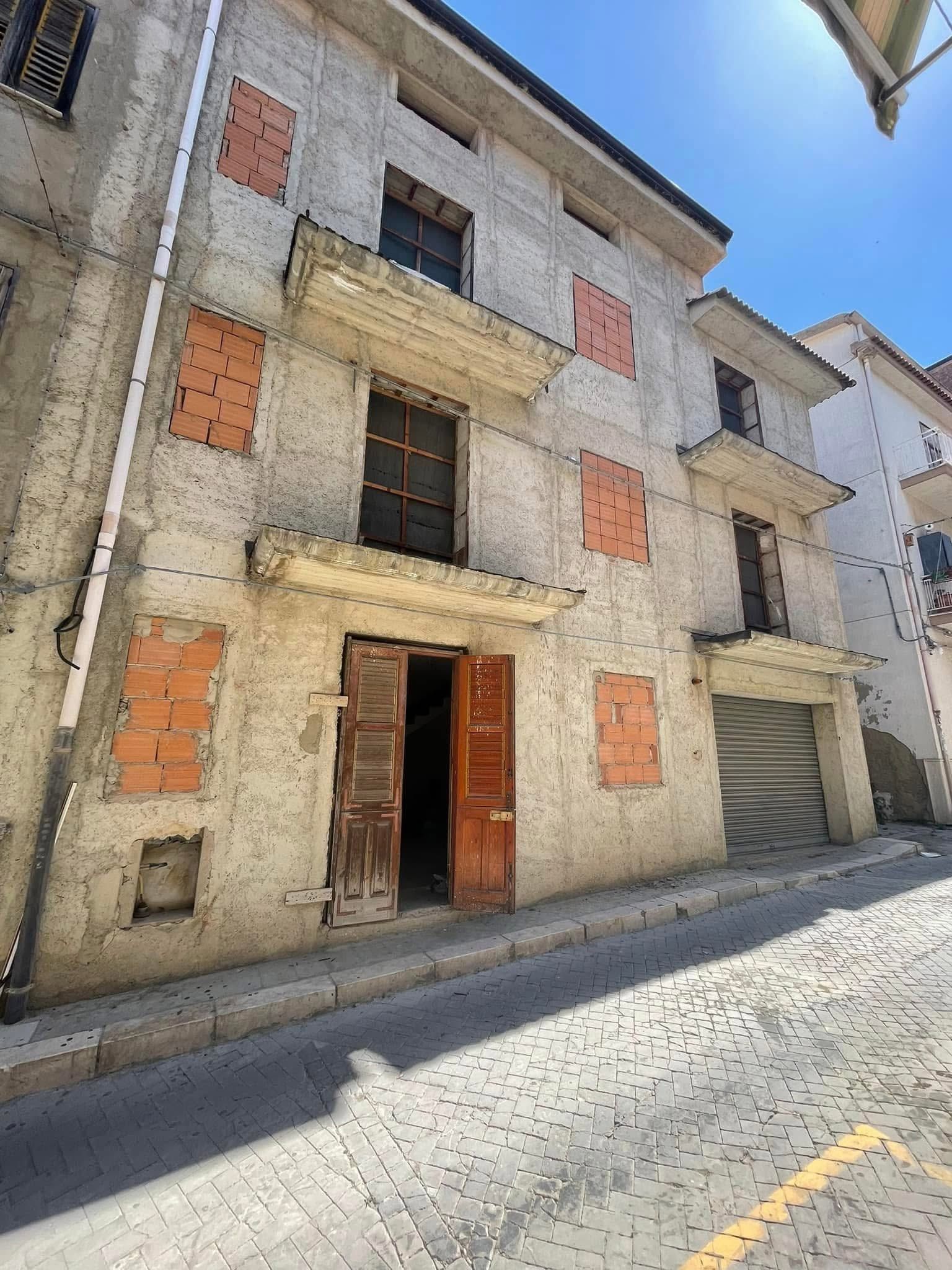 Vecchio edificio incompiuto di tre piani con pannelli in mattoni a vista e porta d'ingresso aperta, situato su una stretta via soleggiata.