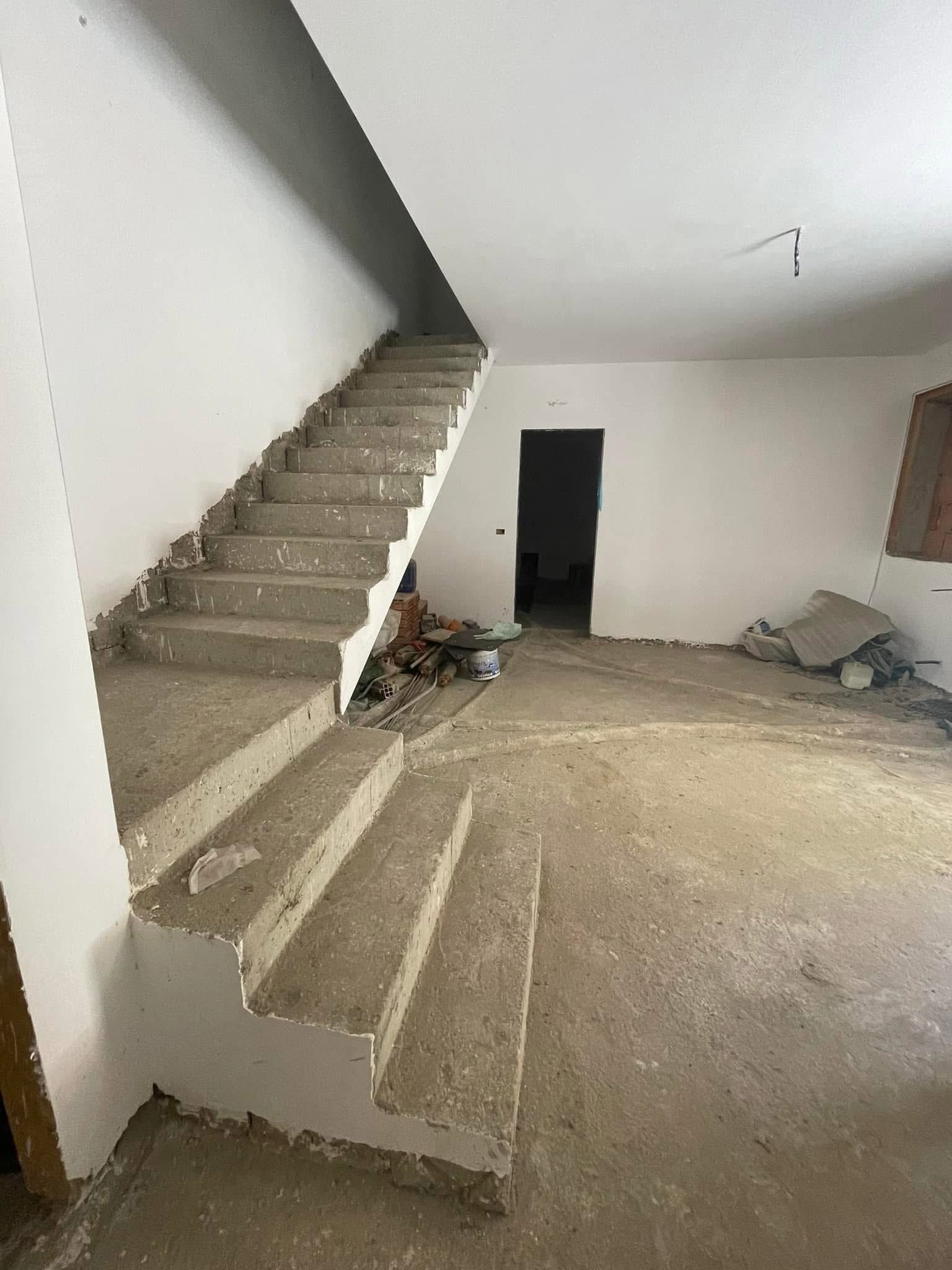 Interno non rifinito con scala in cemento, pareti spoglie e detriti sul pavimento.