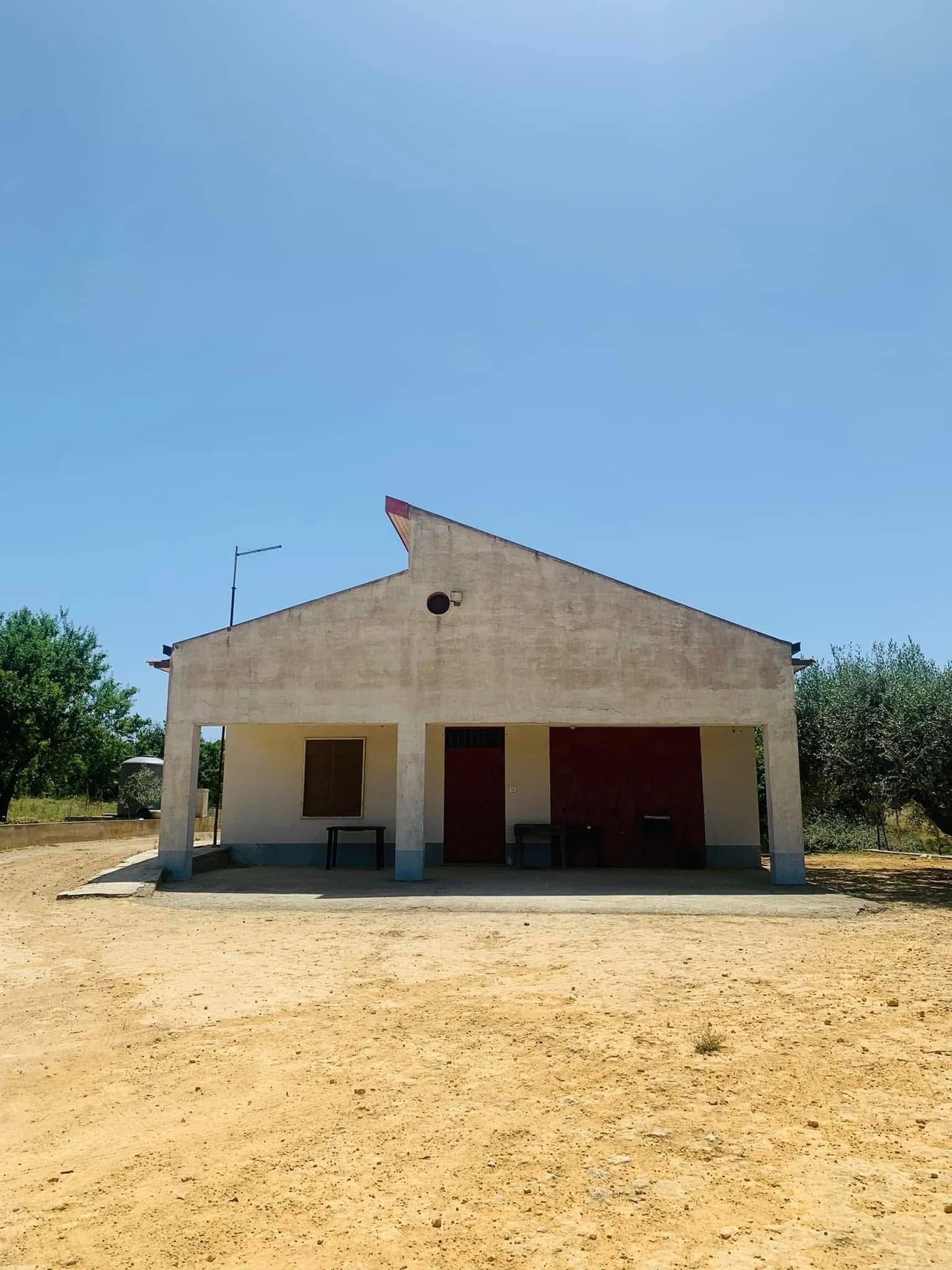 Piccolo edificio beige con un dettaglio rosso sul tetto, situato al centro di un lotto di terra battuta sotto un cielo azzurro e limpido.