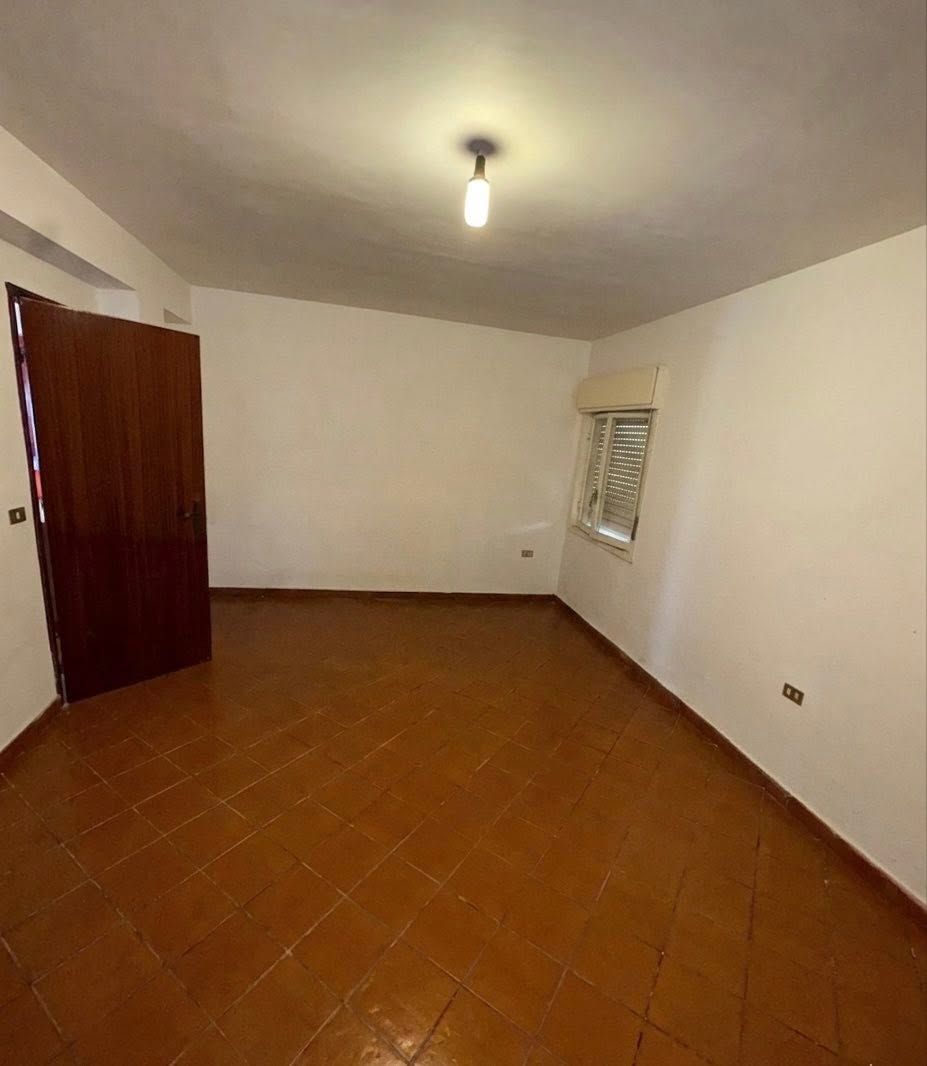 Stanza vuota con pavimento piastrellato, pareti bianche, una porta e una finestra con persiane.