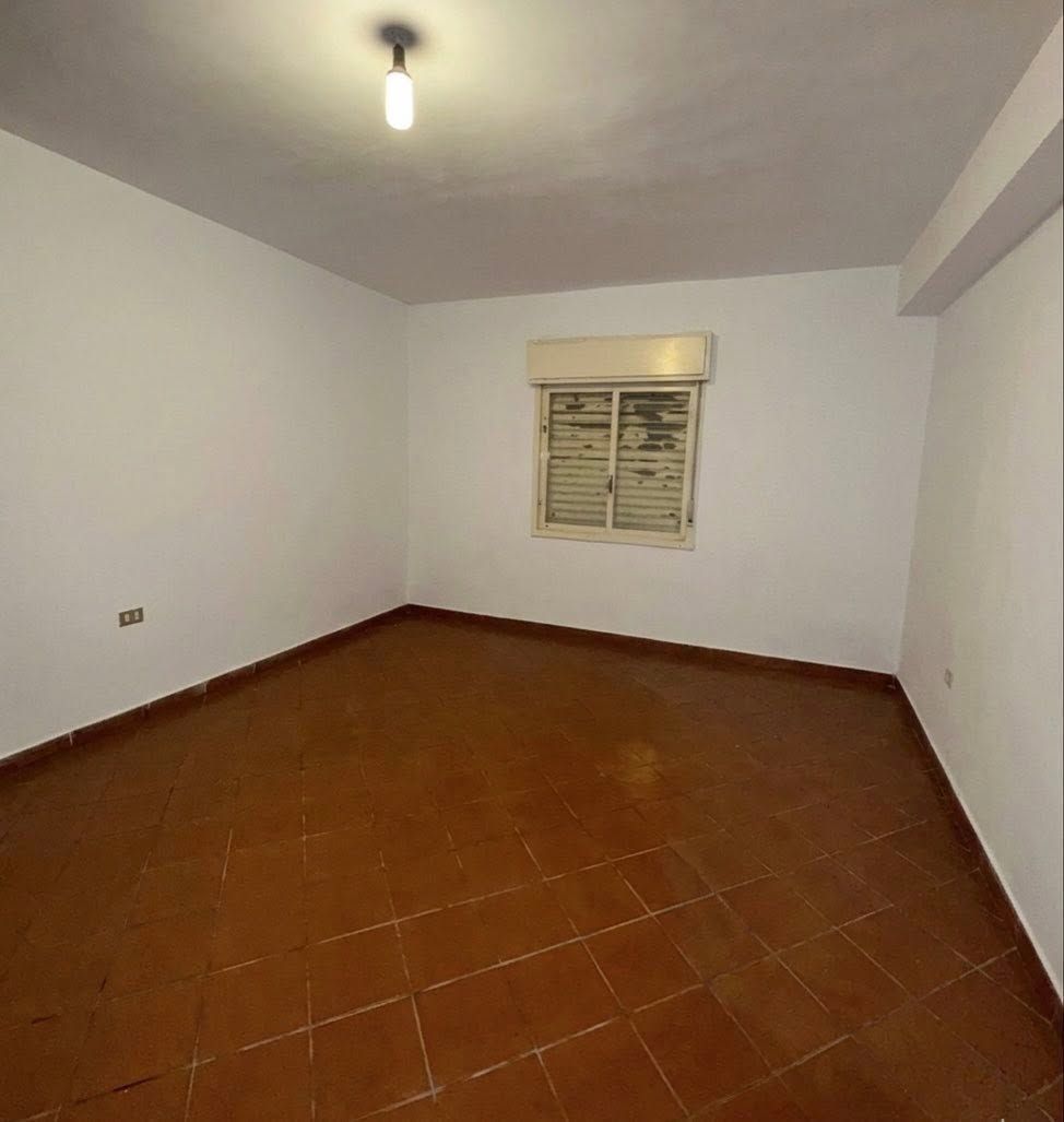 Stanza bianca vuota con pavimento piastrellato di marrone, piccola finestra e un'unica lampada a soffitto.