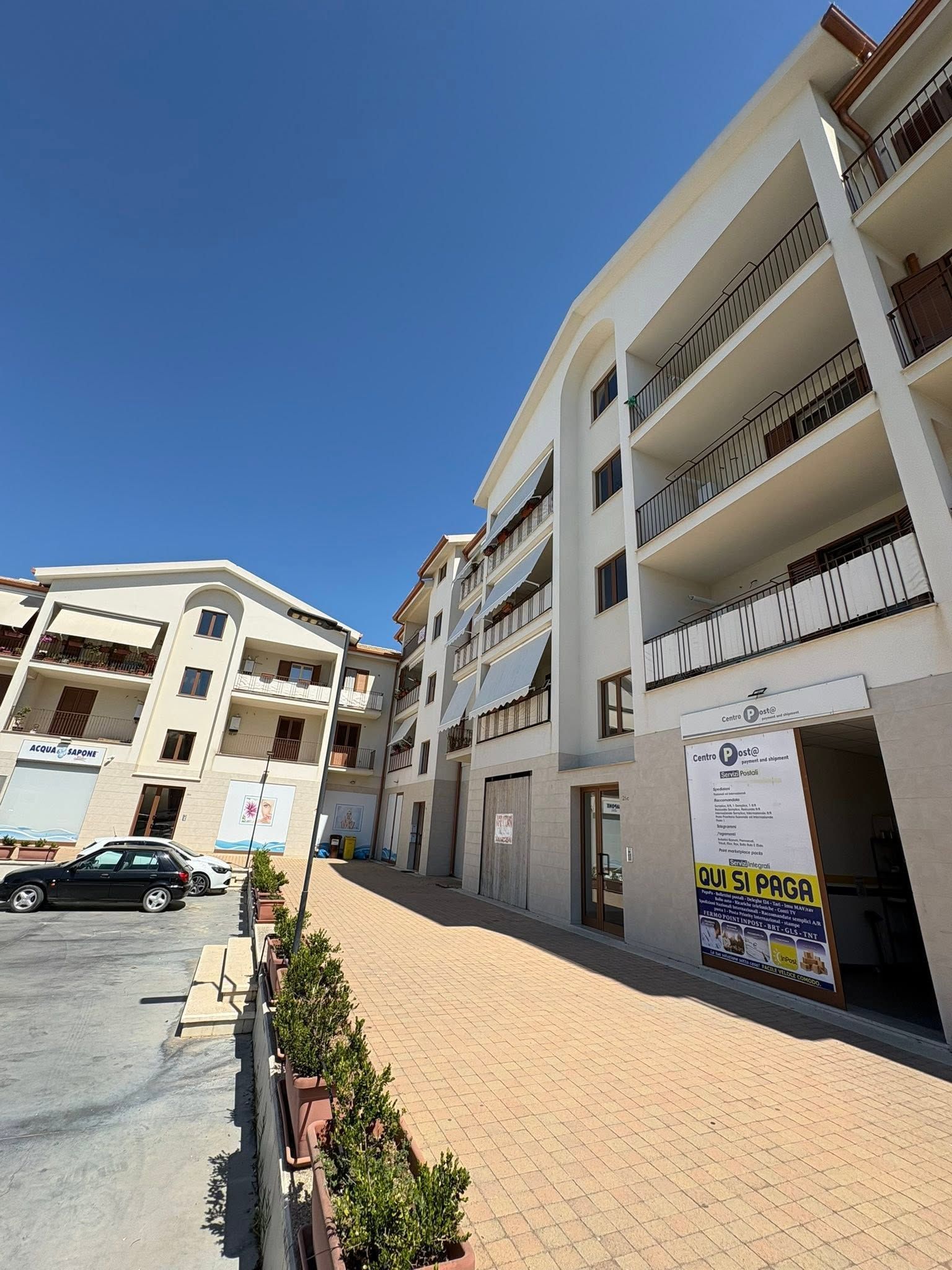 Vista soleggiata di una strada con palazzi bianchi, balconi e un vialetto piastrellato sotto un cielo azzurro.