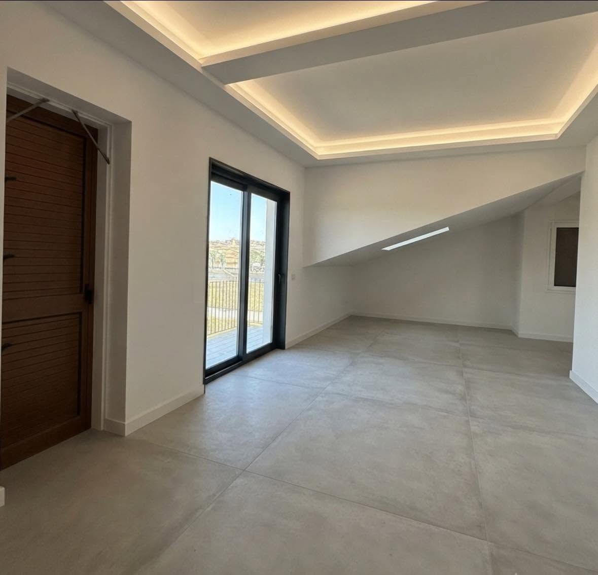 Soggiorno moderno vuoto con pareti bianche, pavimento in piastrelle chiare, faretti a incasso a soffitto e un'ampia porta scorrevole in vetro.