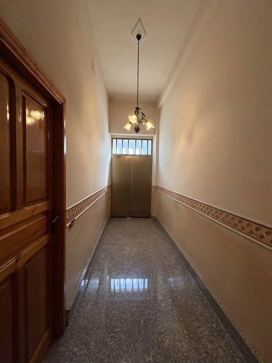 Stretto corridoio con porte in legno, pavimento piastrellato, lampada a soffitto e una porta in vetro smerigliato in fondo.