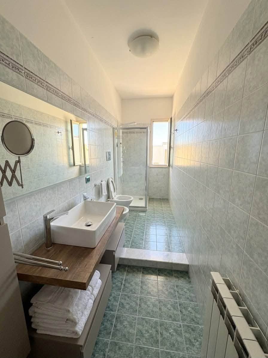 Bagno lungo e stretto con pareti rivestite di piastrelle bianche, pavimento piastrellato di verde, lavandino, specchio e box doccia.