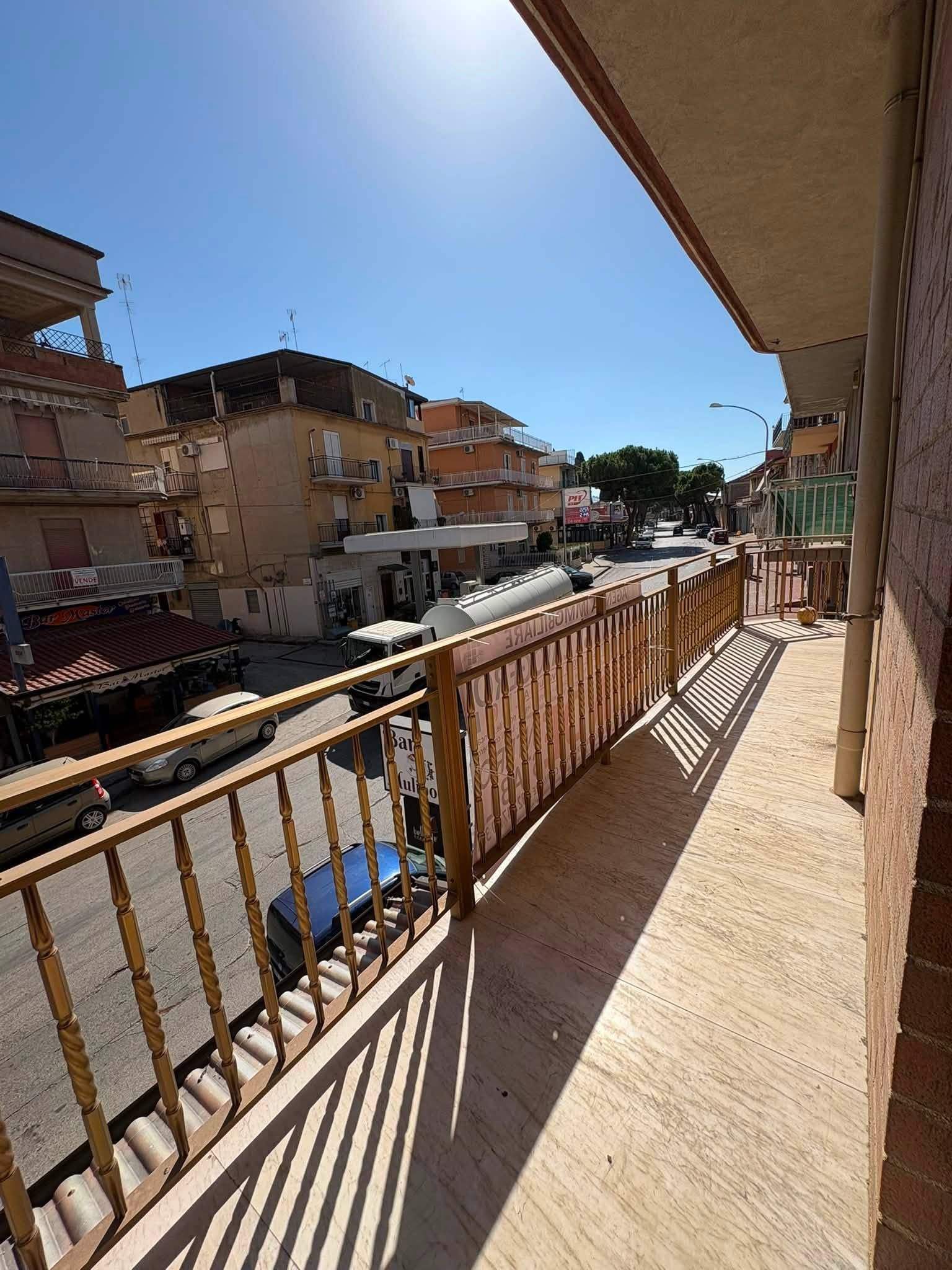 Balcone soleggiato con vista su una strada tranquilla con edifici e auto parcheggiate.