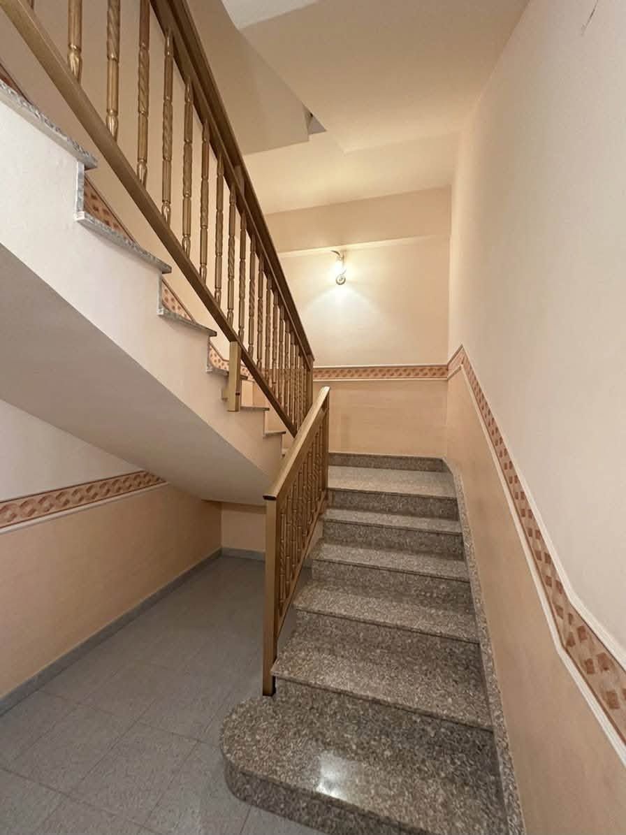Vano scale interno con pareti beige, gradini piastrellati e corrimano in legno illuminati da una luce calda.