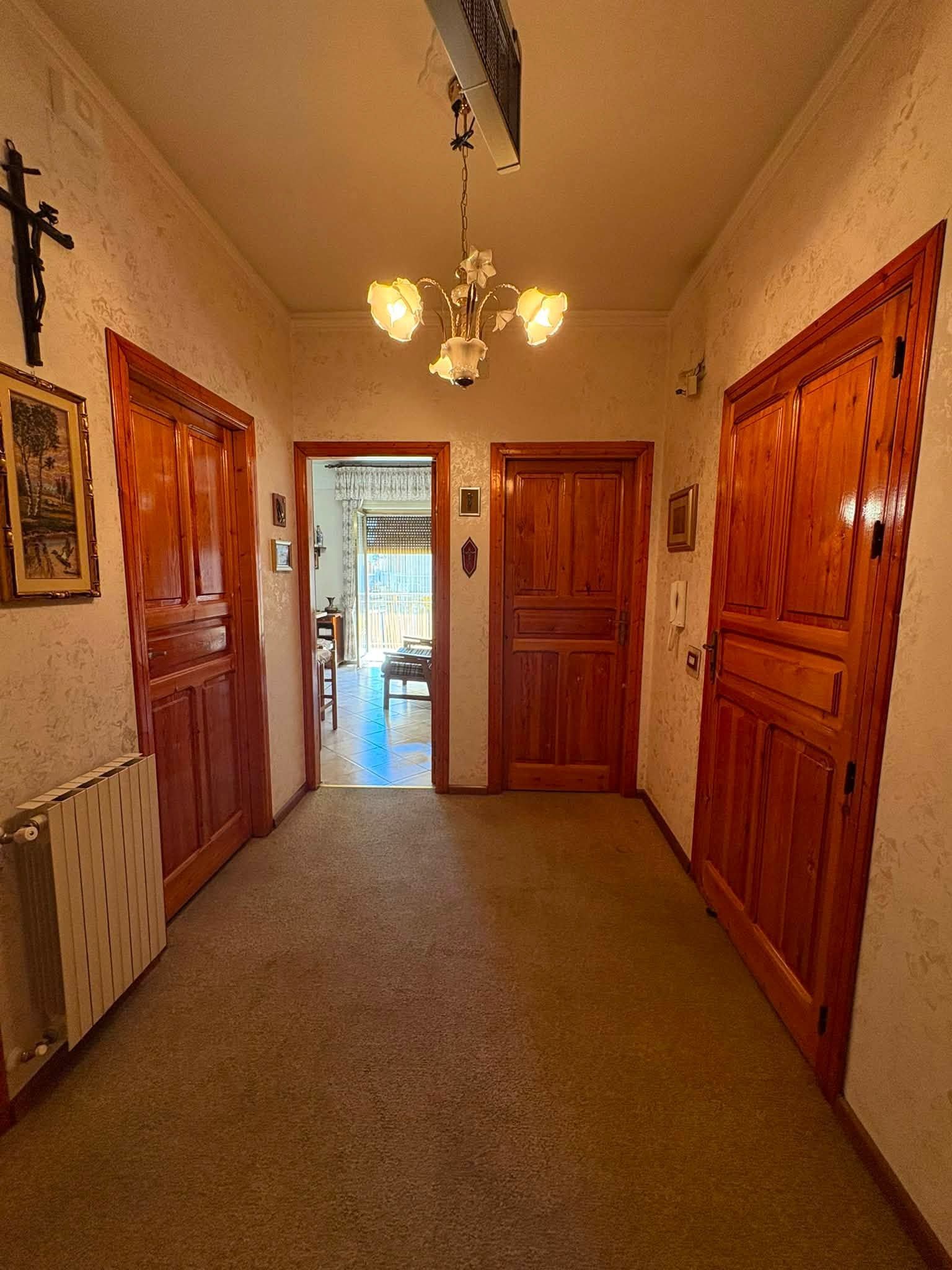 Stretto corridoio con porte in legno, lampadario, termosifone e una stanza luminosa visibile in fondo.
