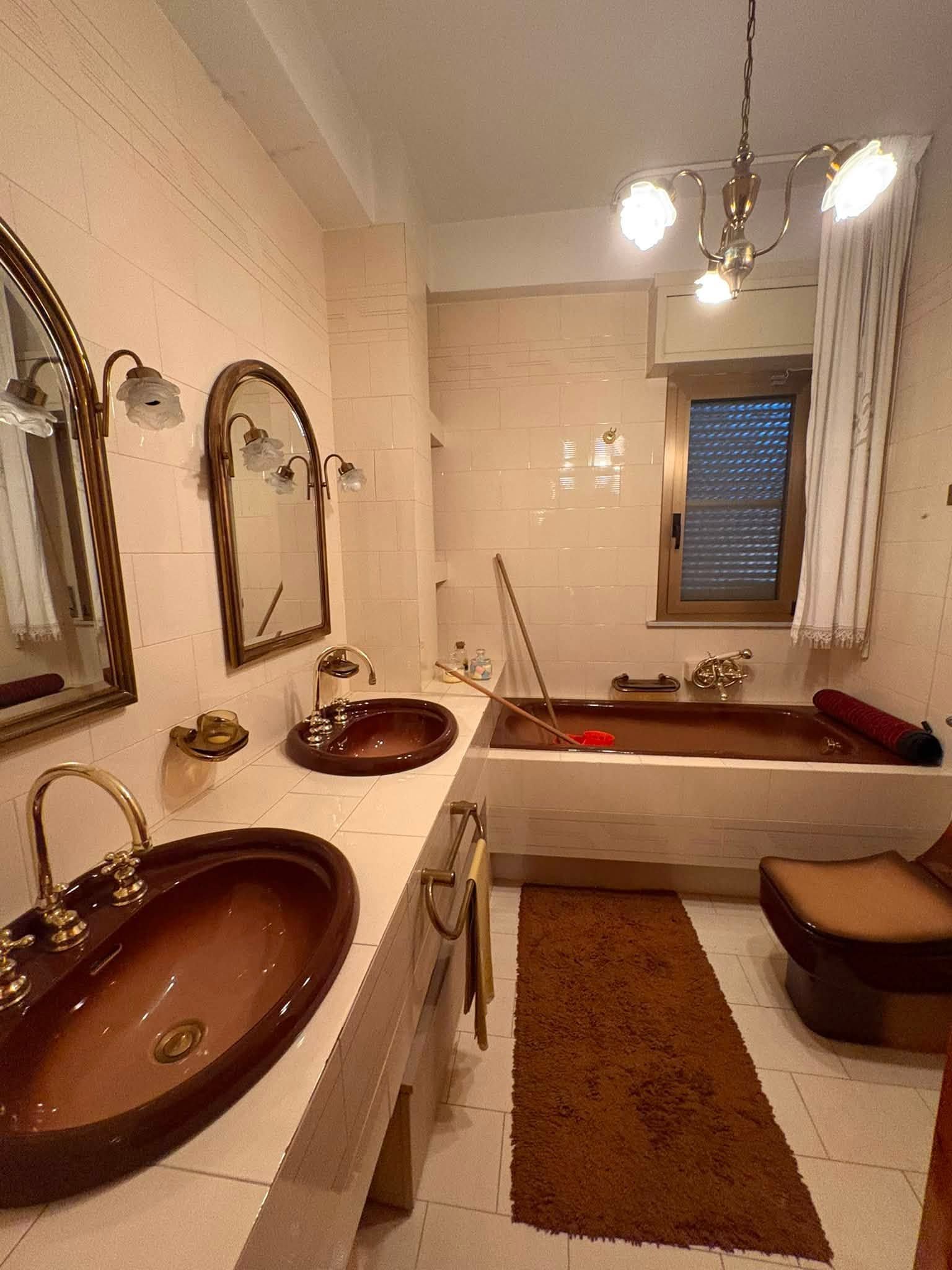 Piccolo bagno con due lavandini, una vasca da bagno, specchi e una finestra con tende.