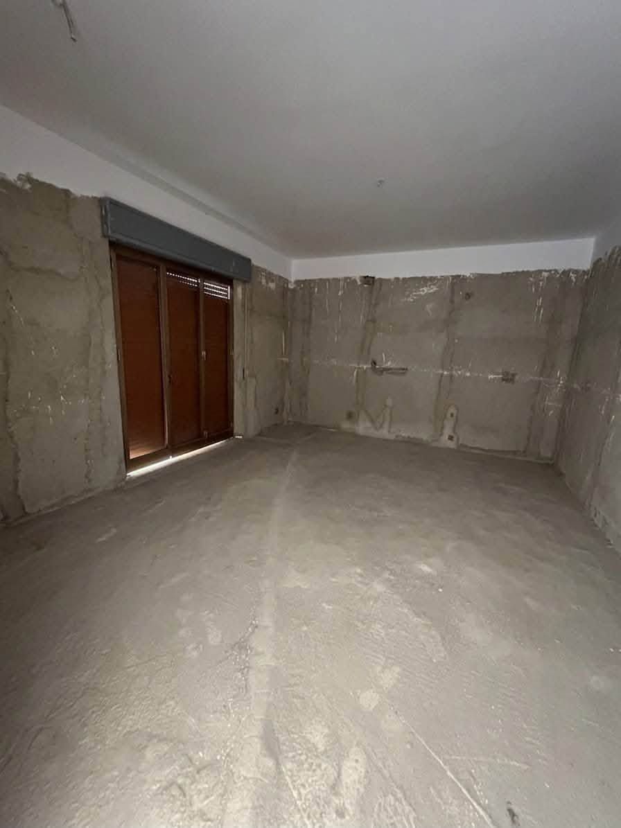 Stanza vuota e non rifinita con pareti e pavimento in cemento grezzo e una persiana marrone.