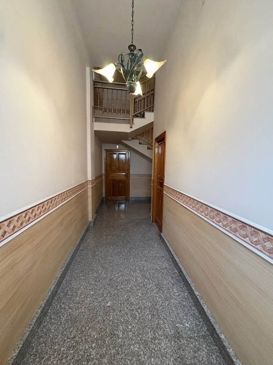Stretto corridoio interno con boiserie decorata, lampadario e scale visibili in fondo.