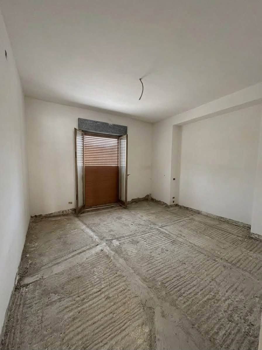 Stanza vuota e non rifinita con pareti bianche, pavimento nudo e una finestra con le persiane chiuse.