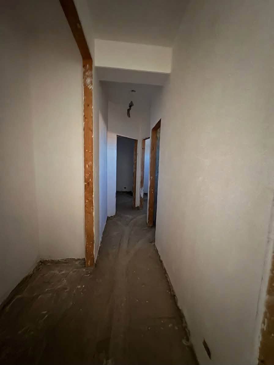 Stretto corridoio non rifinito con pareti bianche, travi in ​​legno a vista e pavimento in cemento.