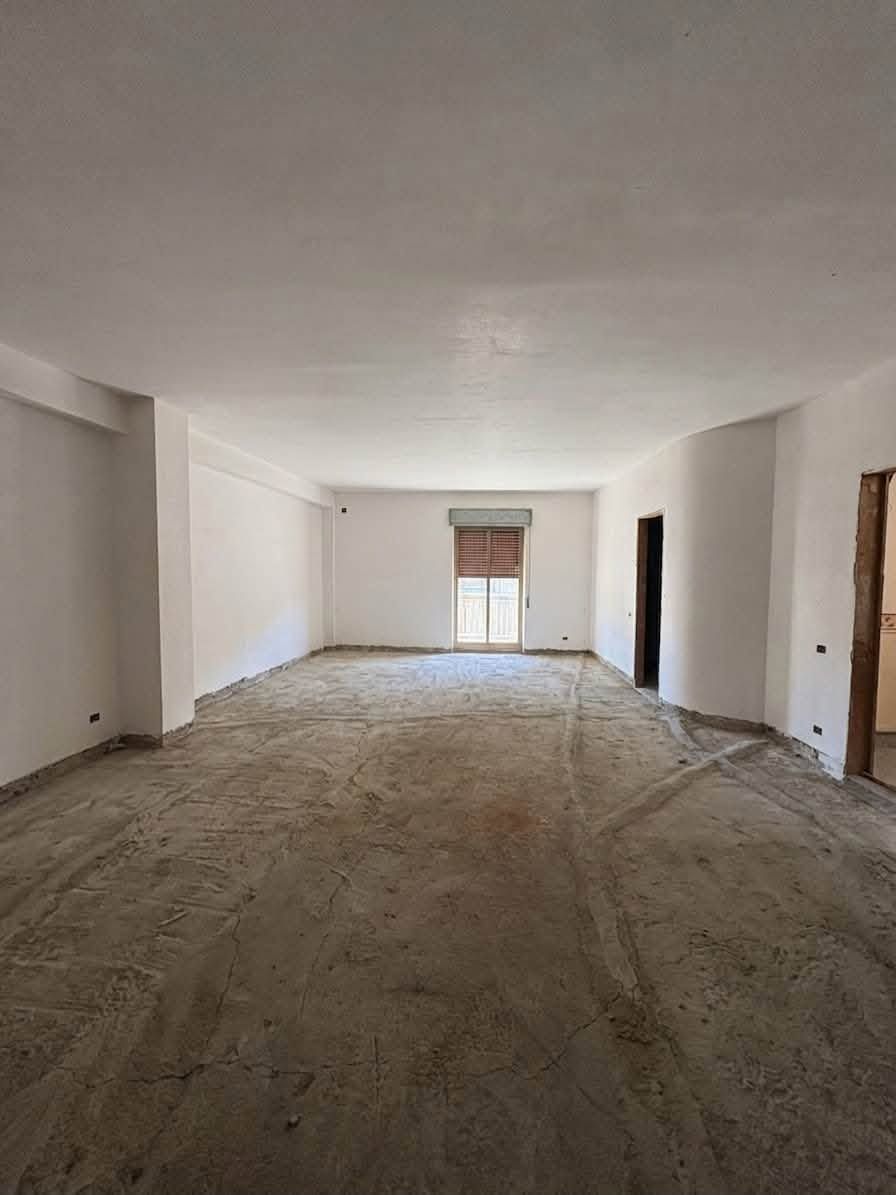 Stanza vuota e spoglia con pareti bianche, pavimento grezzo e una finestra in fondo.