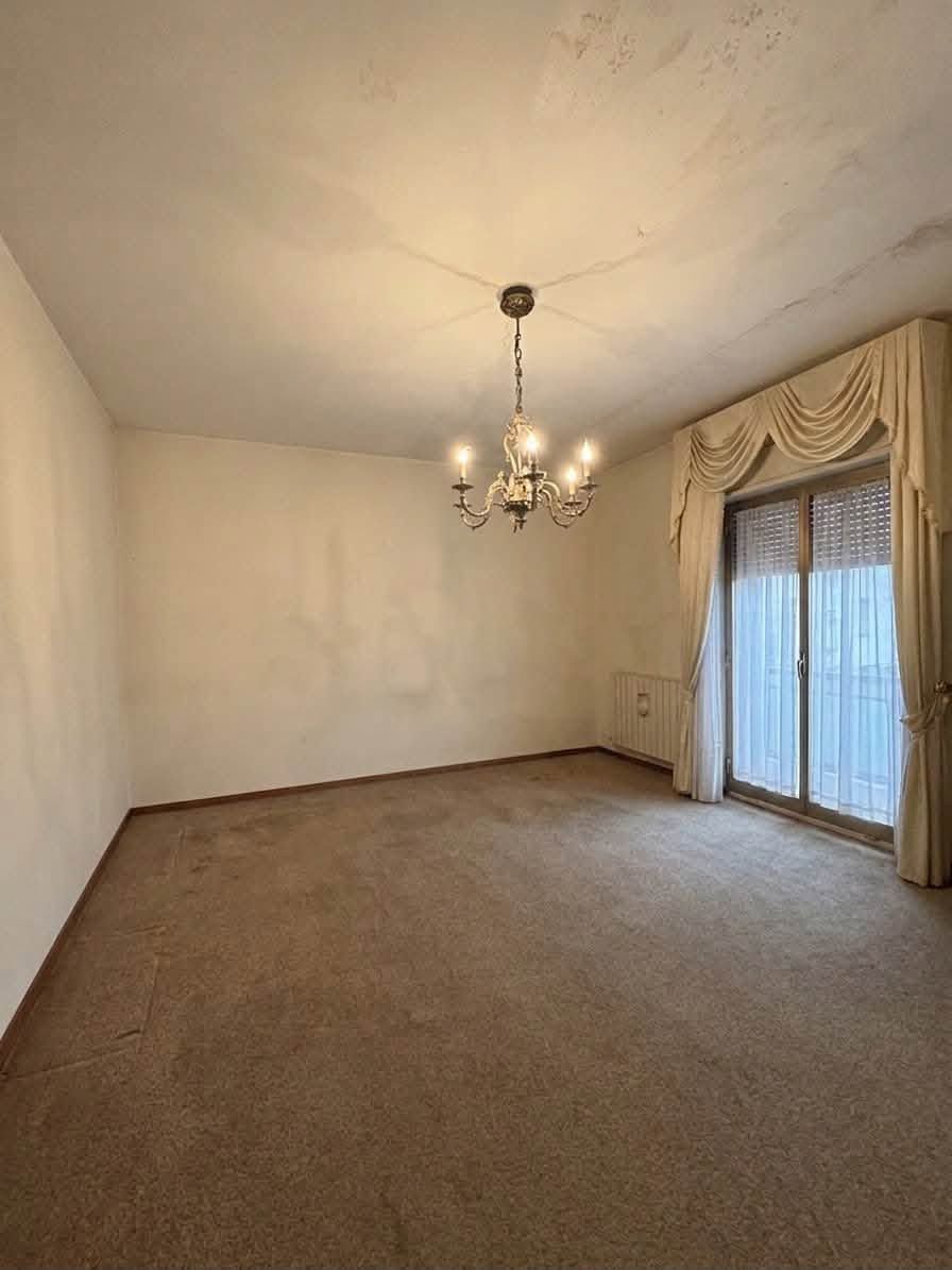 Soggiorno vuoto color beige con lampadario e porta scorrevole in vetro con tende.