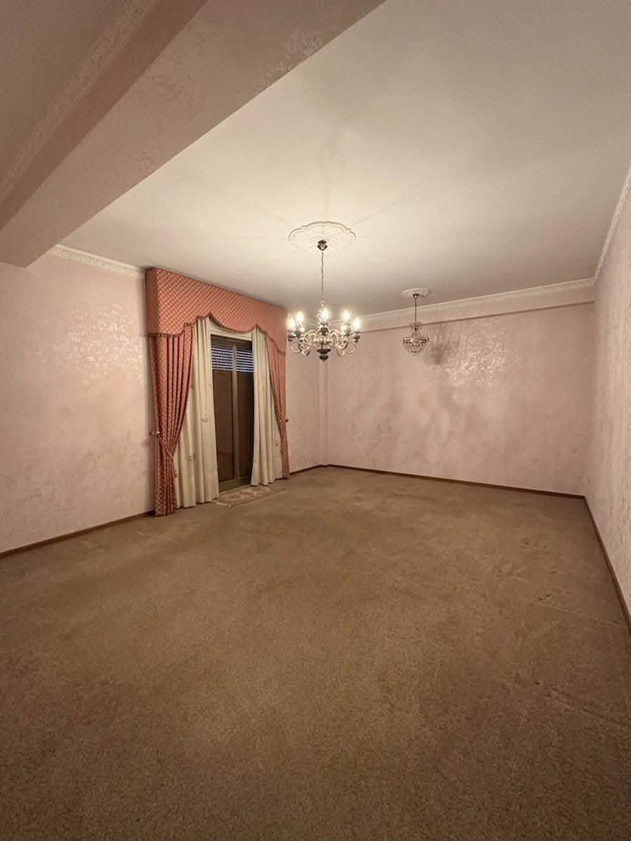 Soggiorno vuoto con moquette rosa, lampadario, carta da parati a motivi e porta drappeggiata.