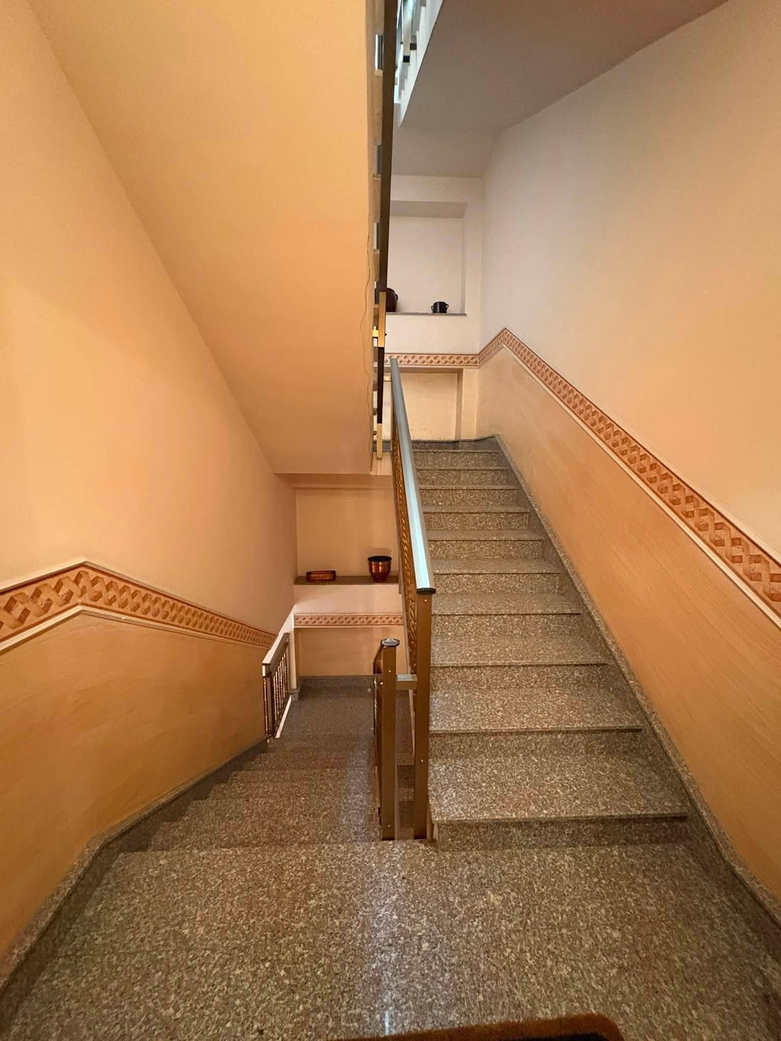 Scala interna stretta con pareti beige, finiture in piastrelle decorate e gradini grigi punteggiati che conducono verso l'alto.