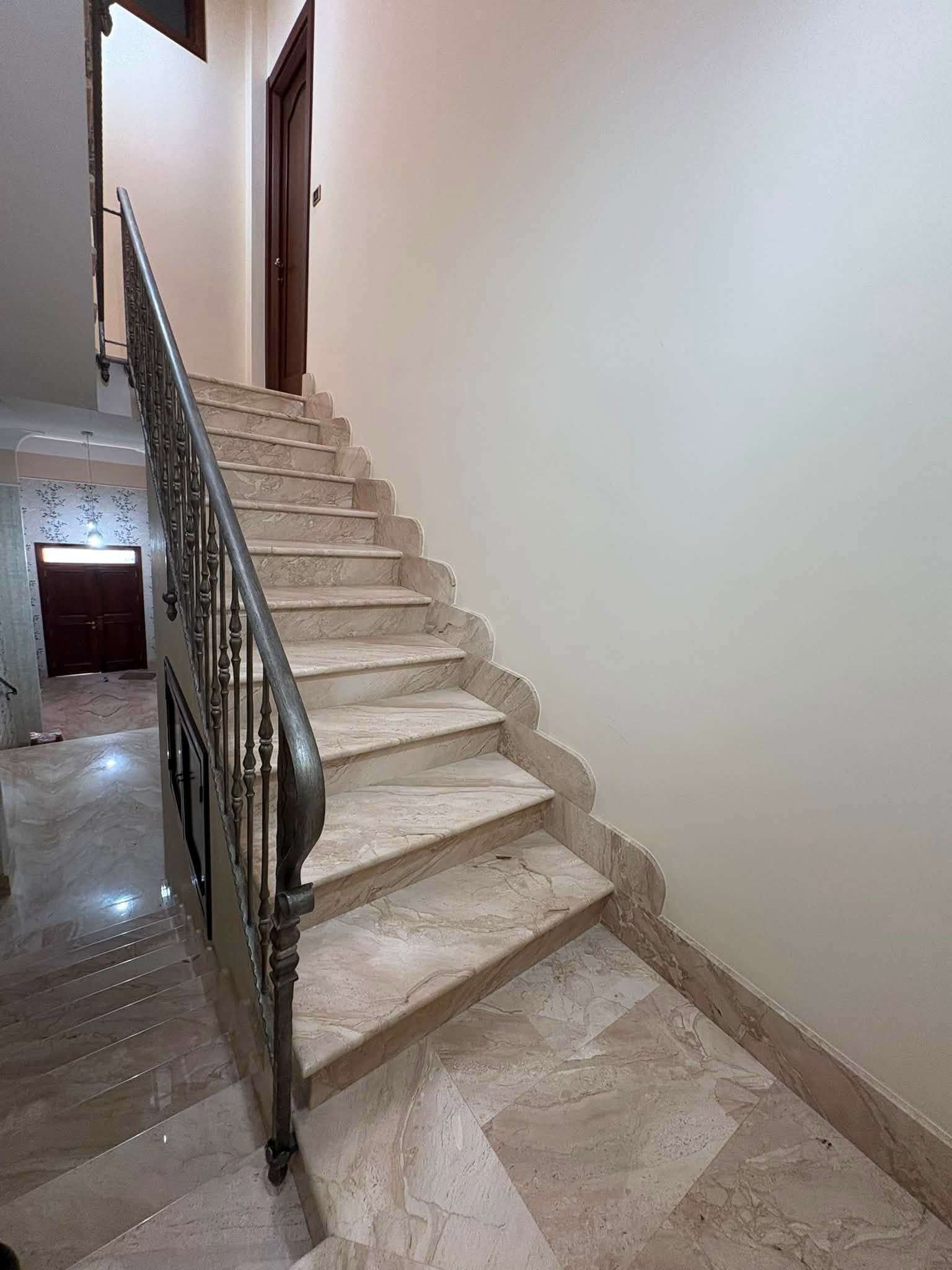 Scala interna con gradini in marmo beige, ringhiera in metallo e pareti bianche in uno stretto corridoio