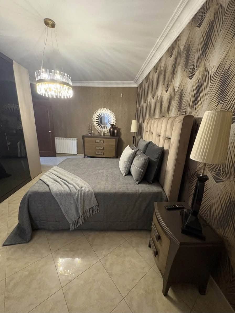 Elegante camera da letto con letto capitonné, biancheria da letto grigia, lampadario, comò con specchio e pavimento piastrellato beige.