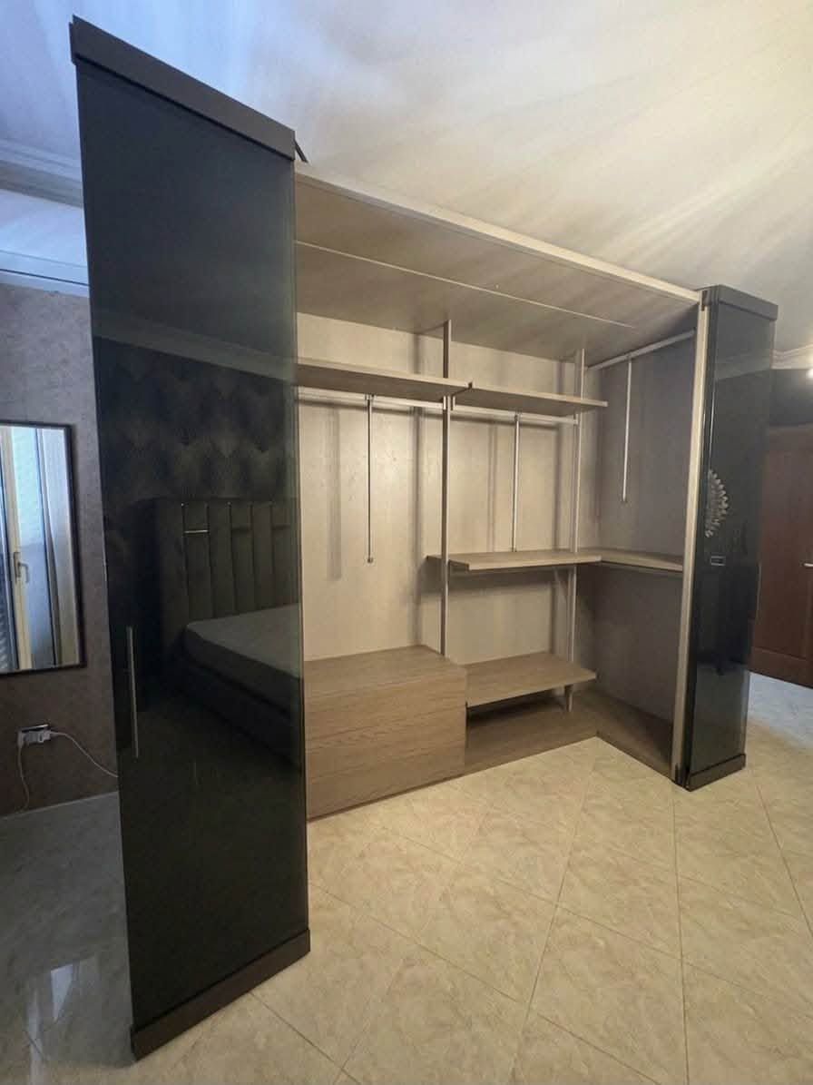 Cabina armadio aperta con ante in vetro con cornice scura, appendiabiti, mensole e panca in una stanza beige.