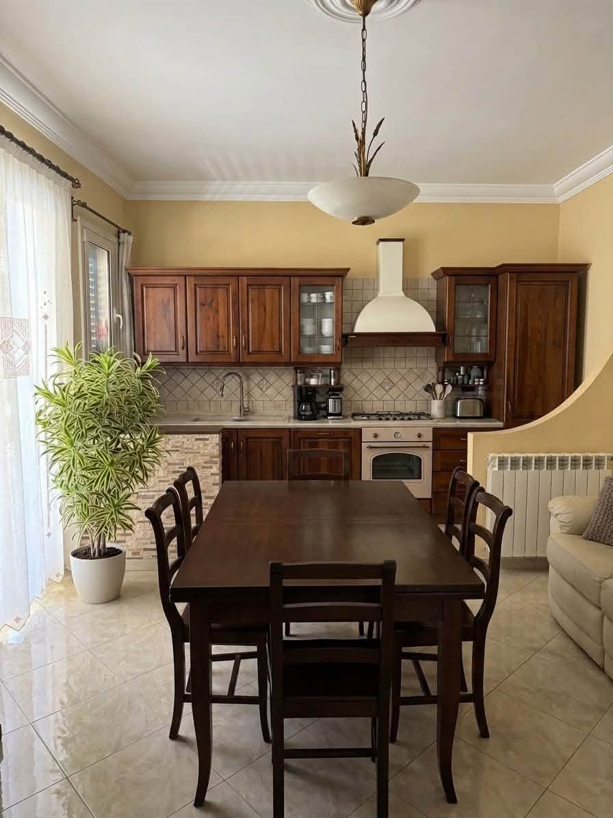 Sala da pranzo e cucina con tavolo in legno, armadi, divano e grande finestra con tende trasparenti.