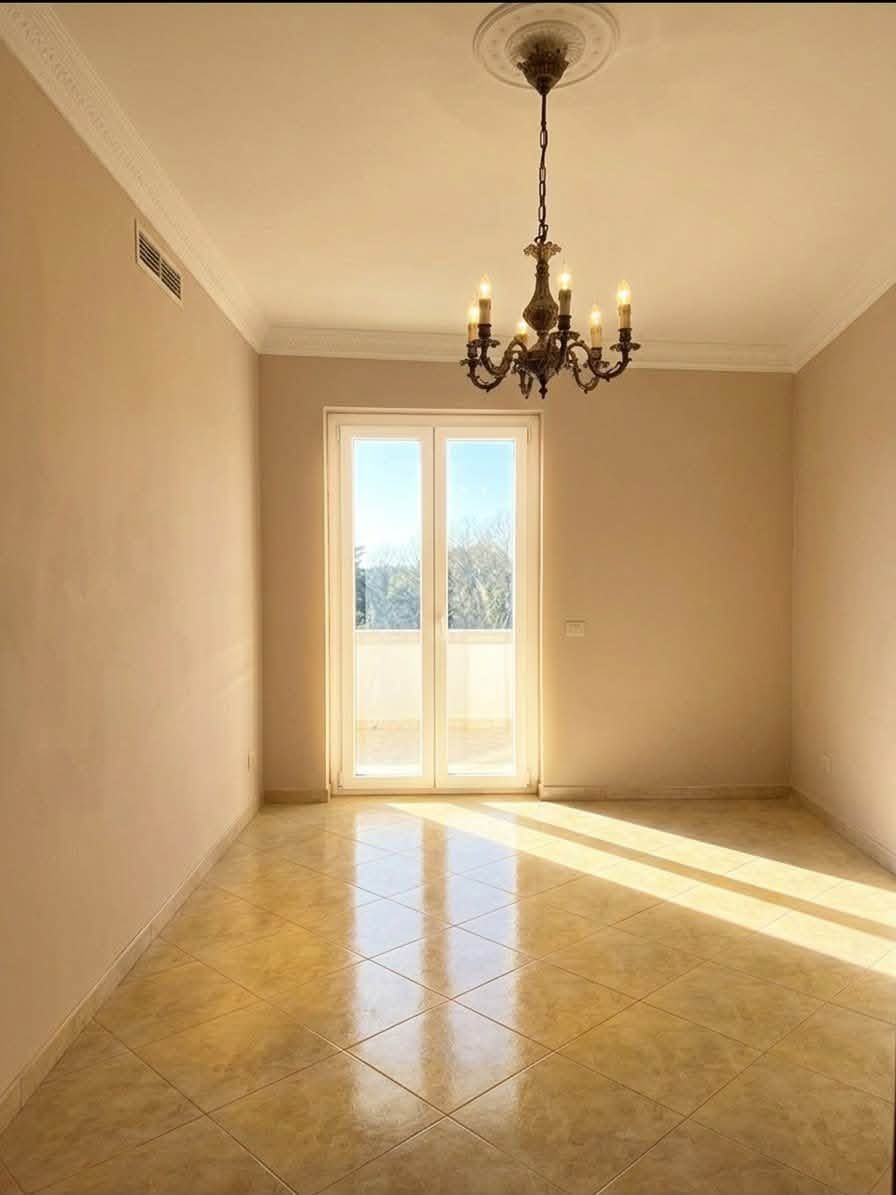 Stanza vuota color beige con lampadario, pavimento piastrellato e luce del sole che filtra attraverso un'alta finestra.