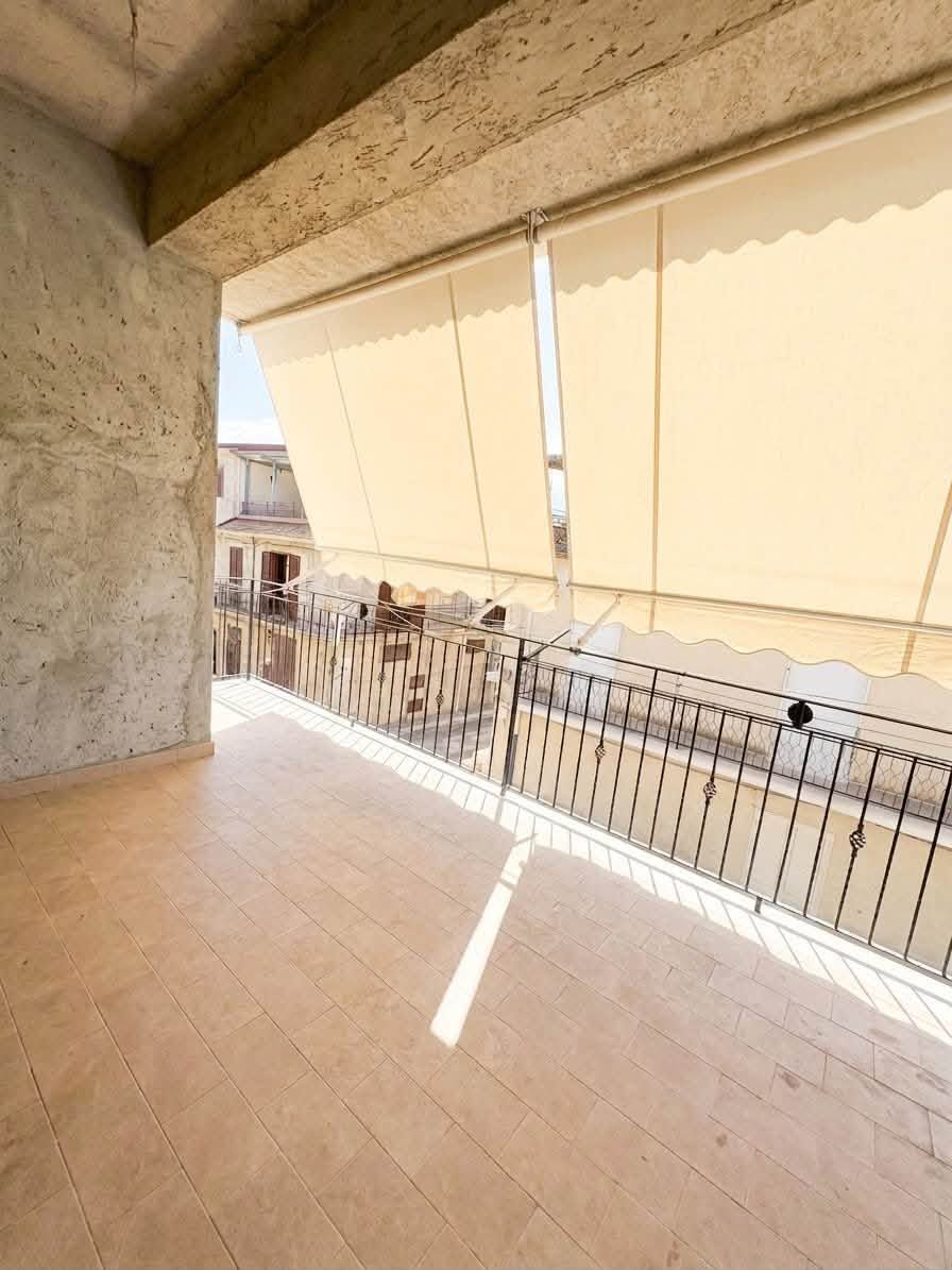 Balcone coperto con pavimento in piastrelle beige, colonne in cemento, ringhiera e tende da sole color crema, affacciato su una strada sottostante.