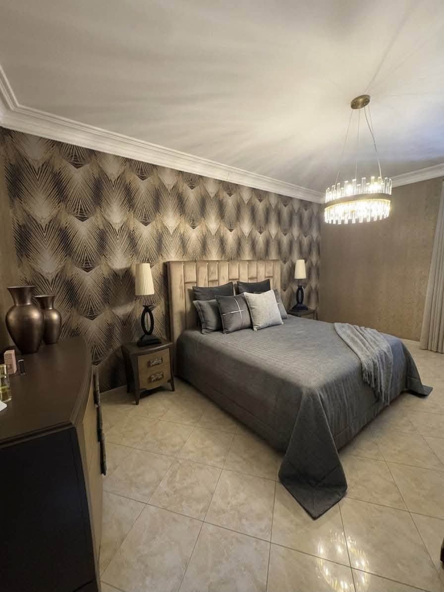 Elegante camera da letto con copriletto grigio, carta da parati a motivi geometrici, lampadario e pavimento piastrellato.