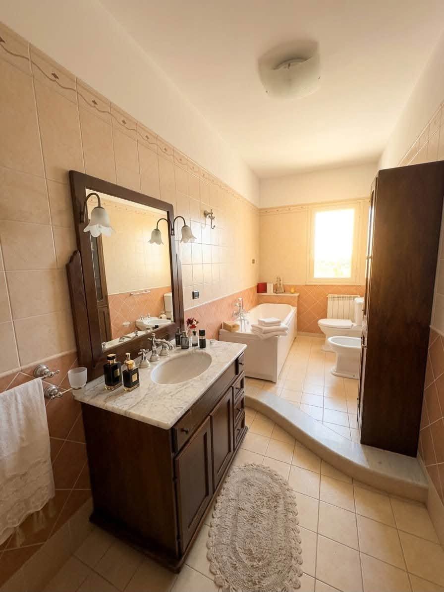 Bagno luminoso con doppio lavabo, ampio specchio, vasca, WC e finestra in fondo.