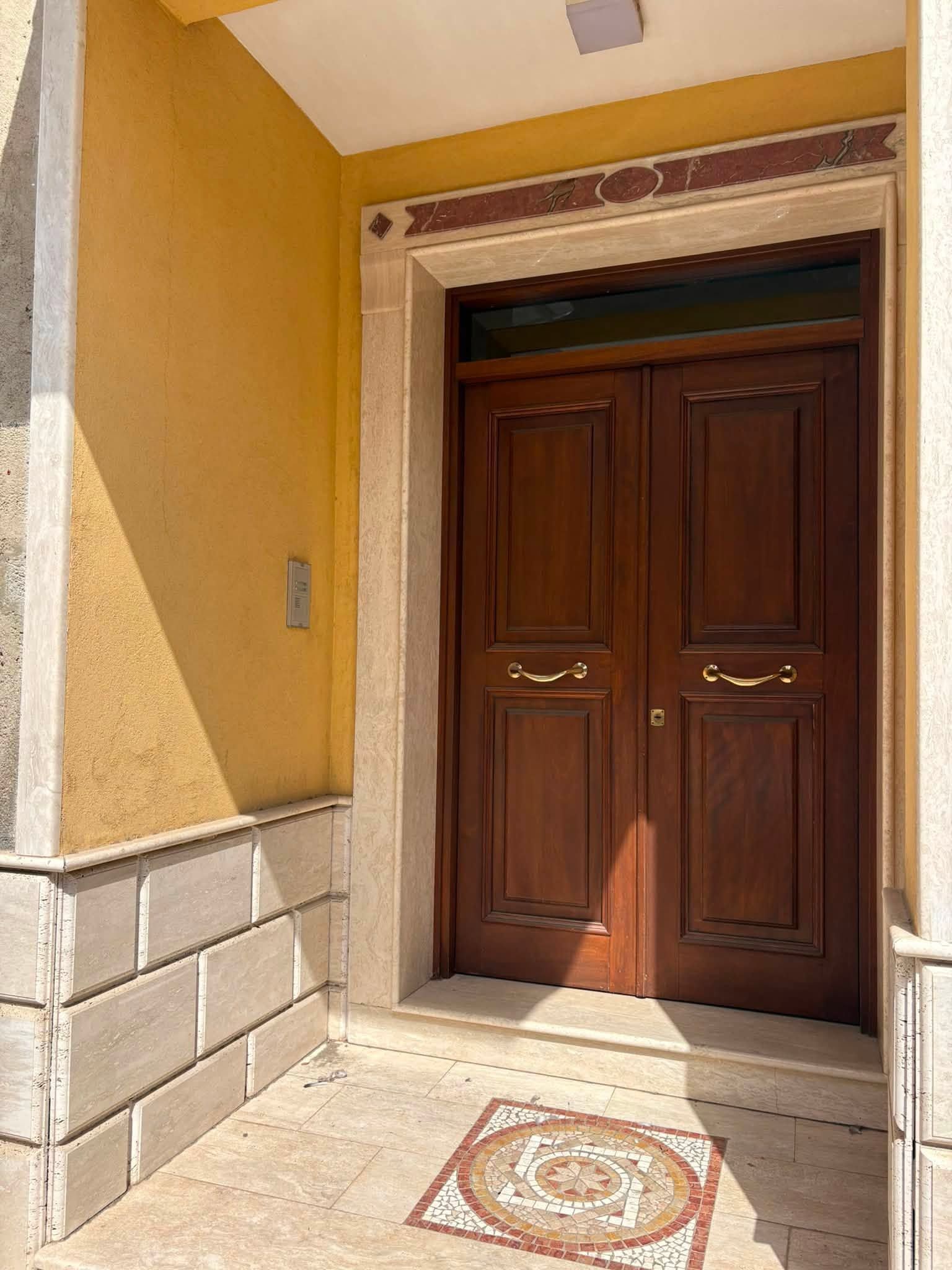 Doppia porta d'ingresso in legno sotto un arco beige con pareti piastrellate e un medaglione decorativo sul pavimento.
