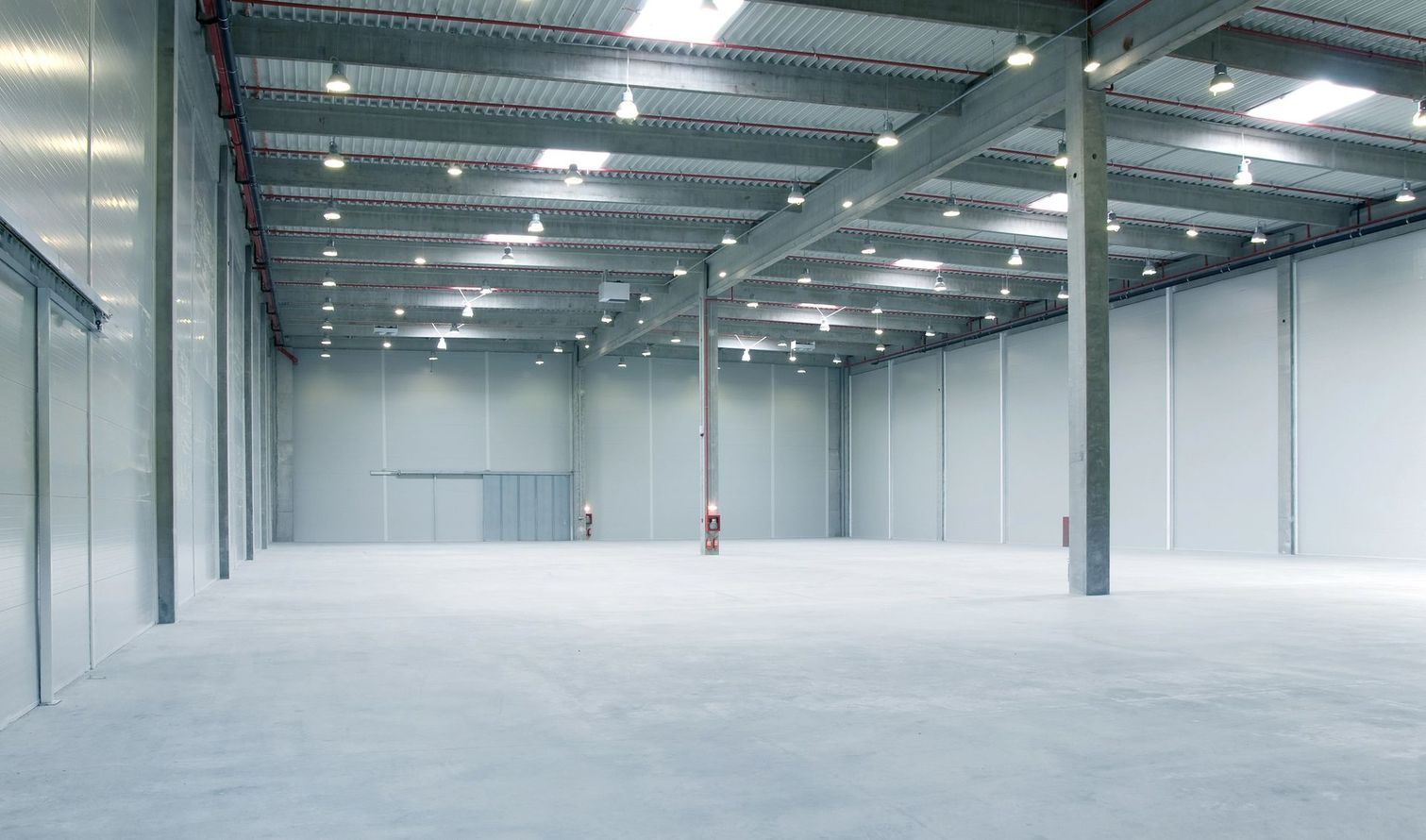 Interno vuoto di un magazzino industriale con pareti bianche, pavimento in cemento e luci a soffitto.