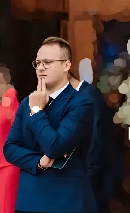 Un uomo in abito blu scuro e occhiali è ritratto pensieroso con la mano sul mento, sullo sfondo sfocato di un paesaggio esterno.