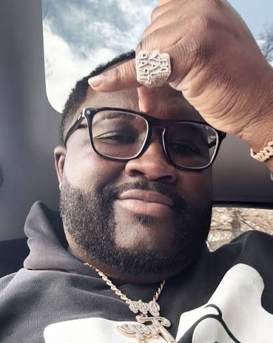 Smiling man wearing glasses and jewelry, resting his hand on his forehead inside a car.